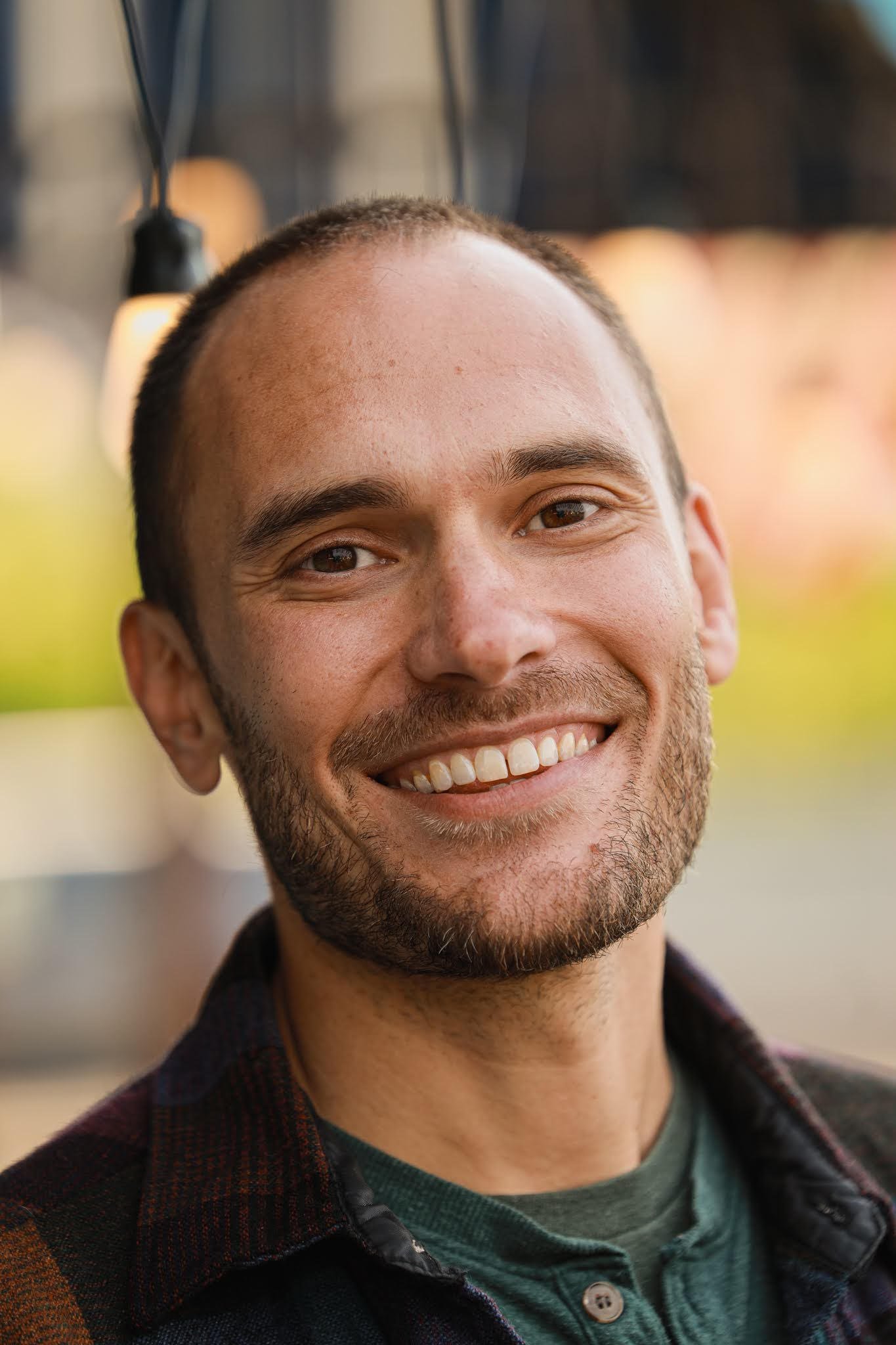 Close-up of smiling young man with short hair and beard, wearing a plaid shirt and green t-shirt, outdoors in natural light.