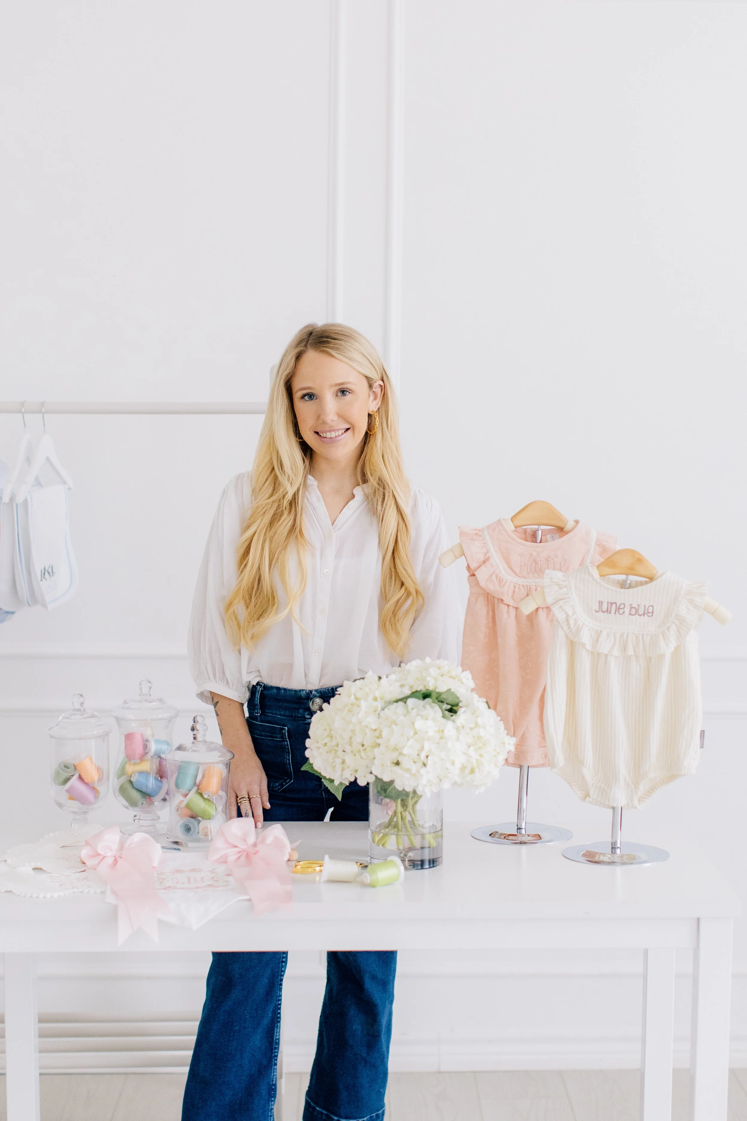 A woman smiles beside a table with white flowers and pink baby clothes on wooden hangers.