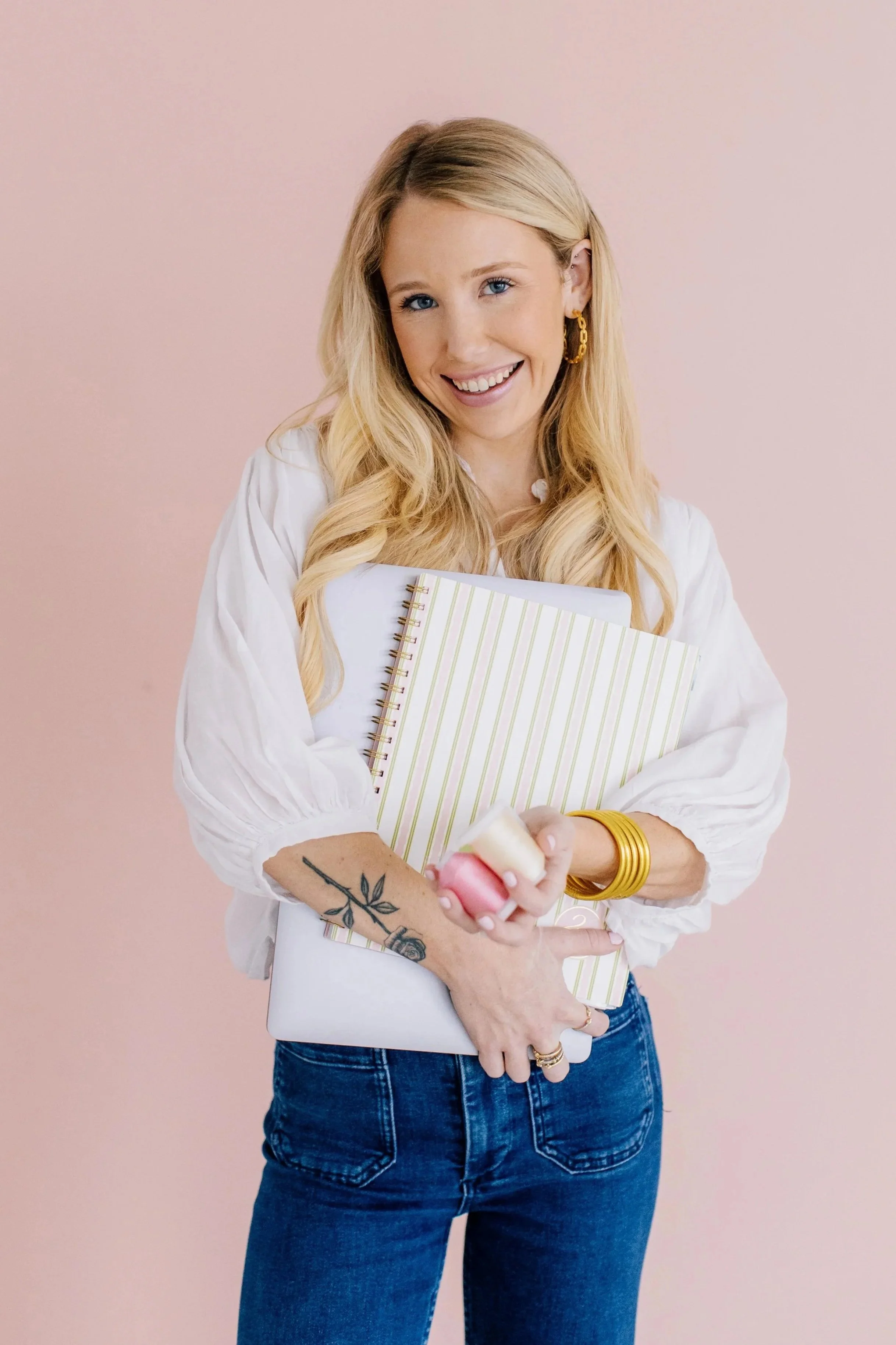 A smiling woman with long blonde hair holding a notebook and pink thread.