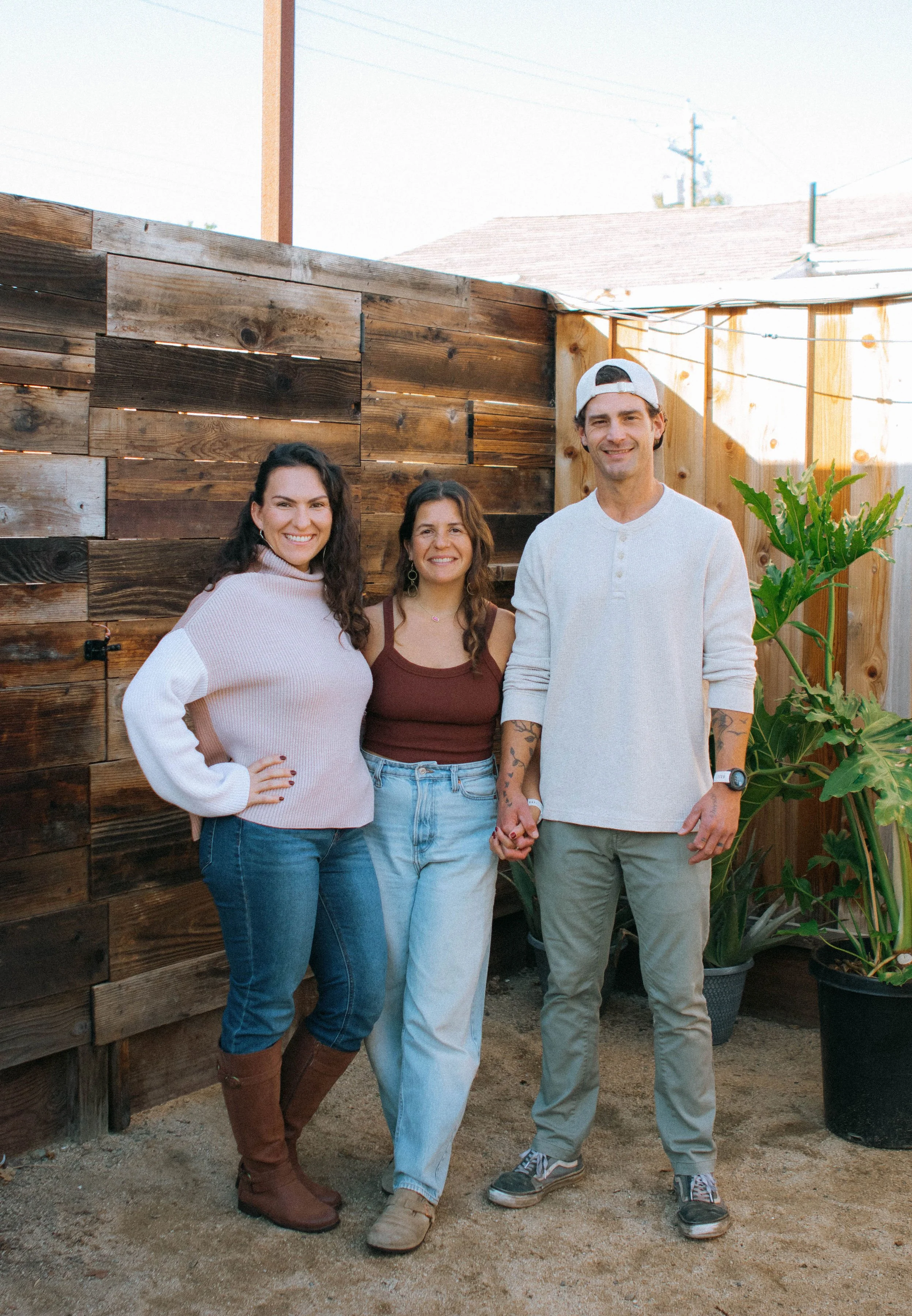 Three people standing outdoors in a backyard with a wooden fence, holding hands and smiling.