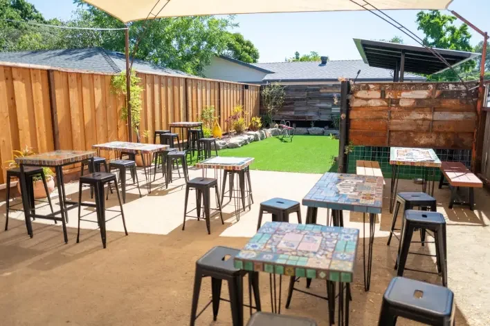 An outdoor patio with colorful tiled tables and black stools, a small grassy area, potted plants, and a wooden fence, under a shade sail.