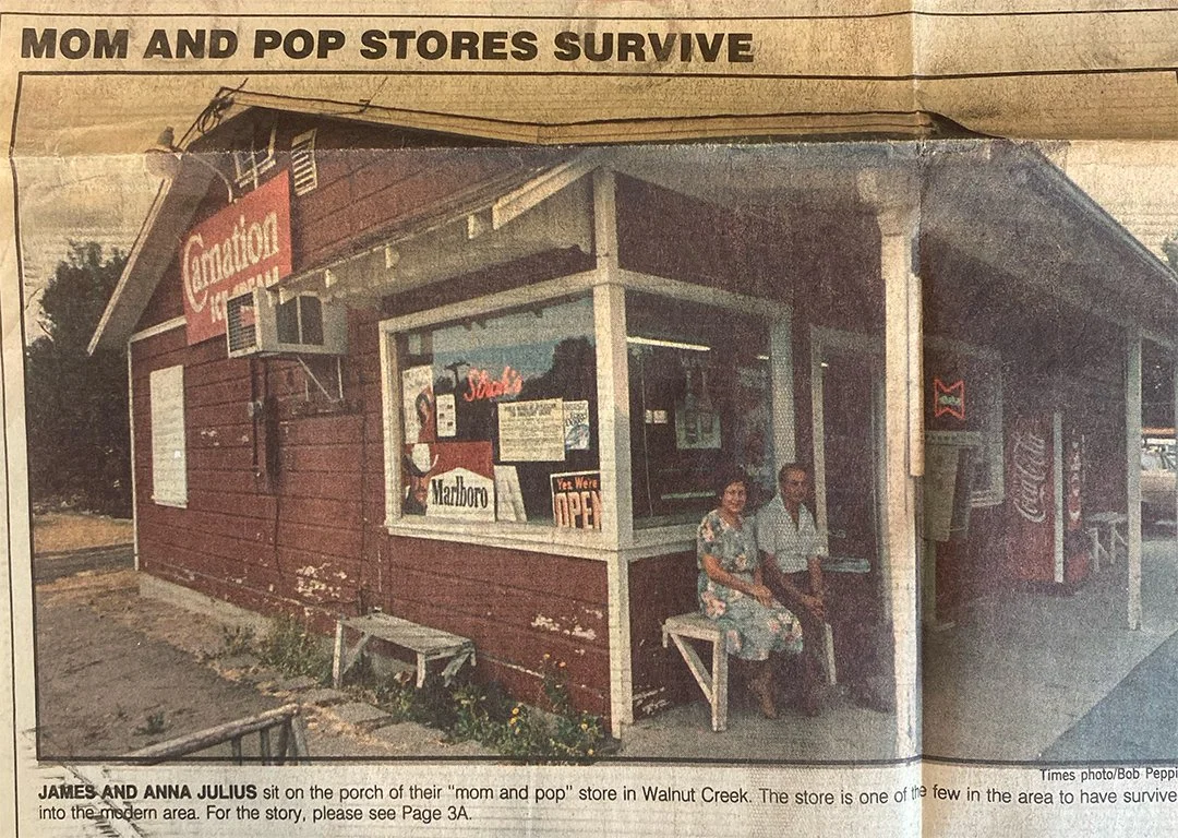 A small red store in Walnut Creek with two people sitting outside on the porch, one woman and one man, with signs for cigarettes and soda in the window.