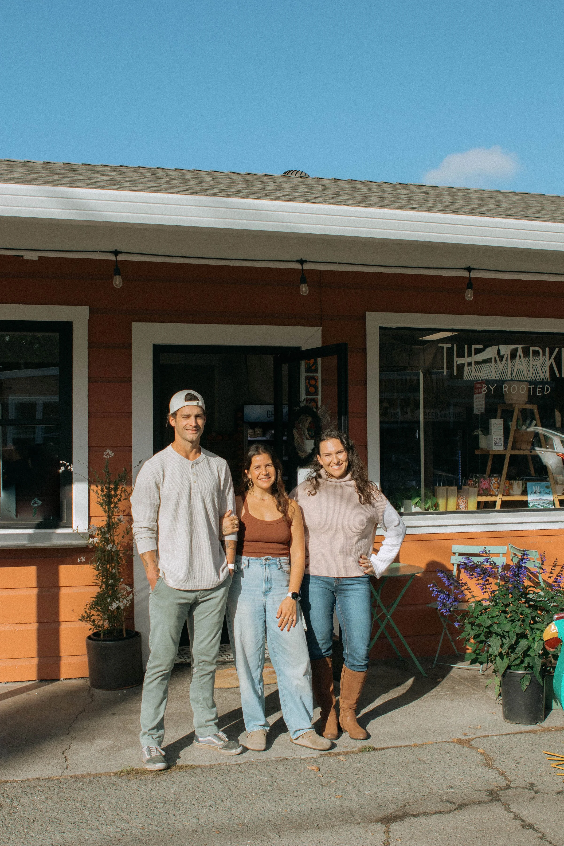Three people standing outside a storefront with plants and chairs in front, smiling at the camera.