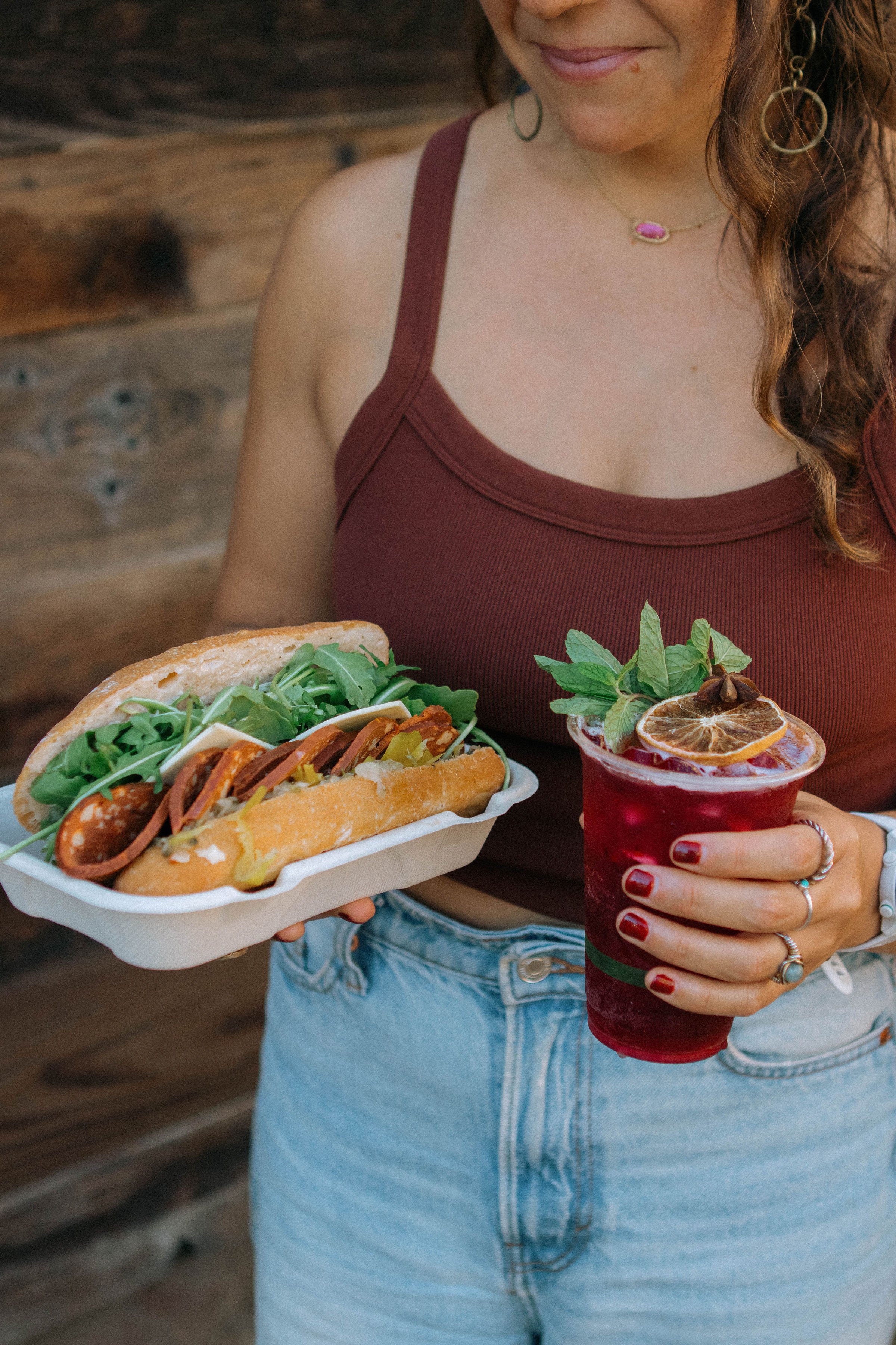A woman wearing a maroon tank top and light blue jeans holding a hot dog topped with various toppings in a foam tray in one hand and a red beverage garnished with dried lemon slices, mint leaves, and star anise in the other hand.
