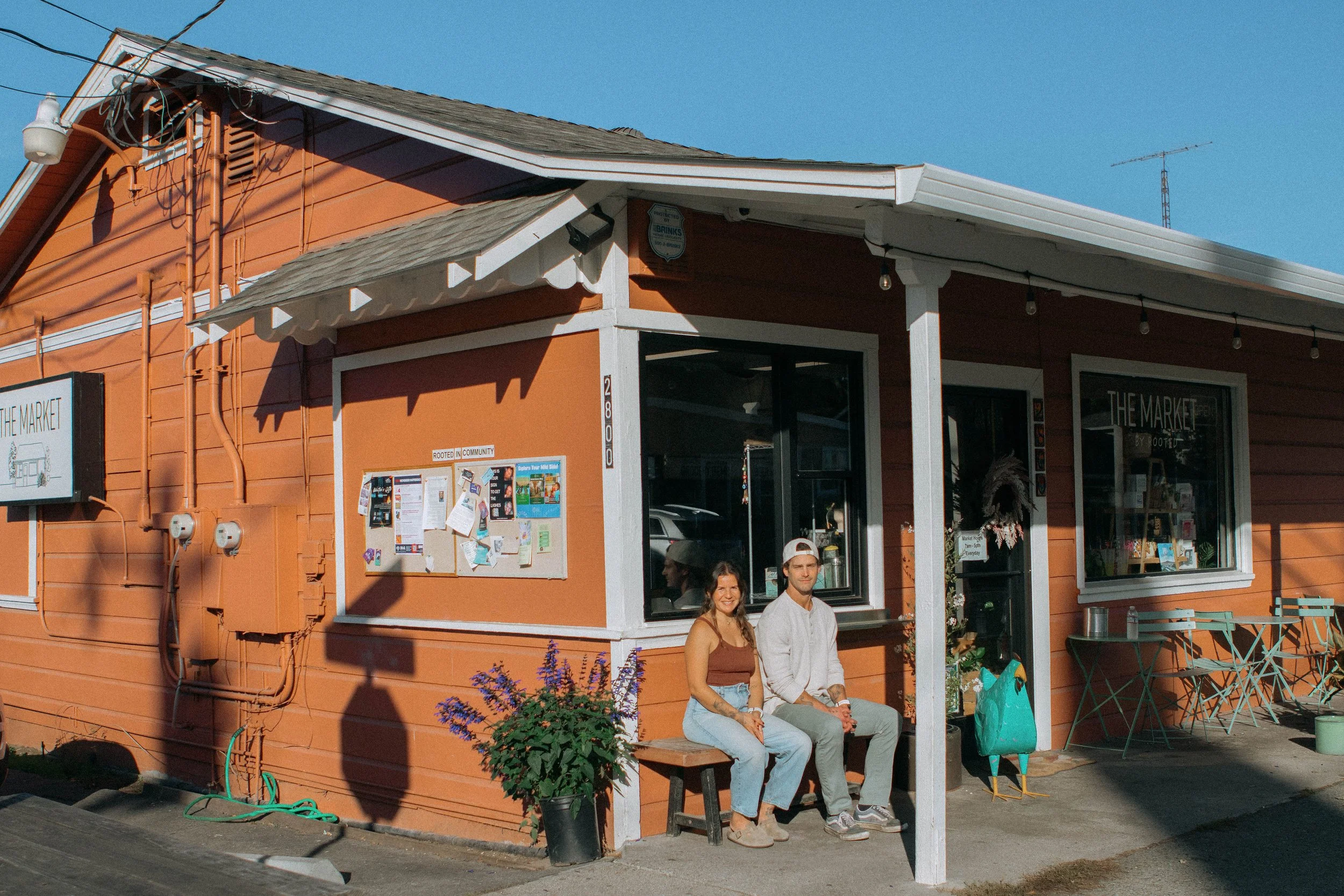A small orange building with white trim houses a shop called 'The Market by Boots'. Two people sit on a bench outside, smiling; one woman and one man. There are potted plants, a bulletin board, and outdoor tables with chairs on the sidewalk in front.