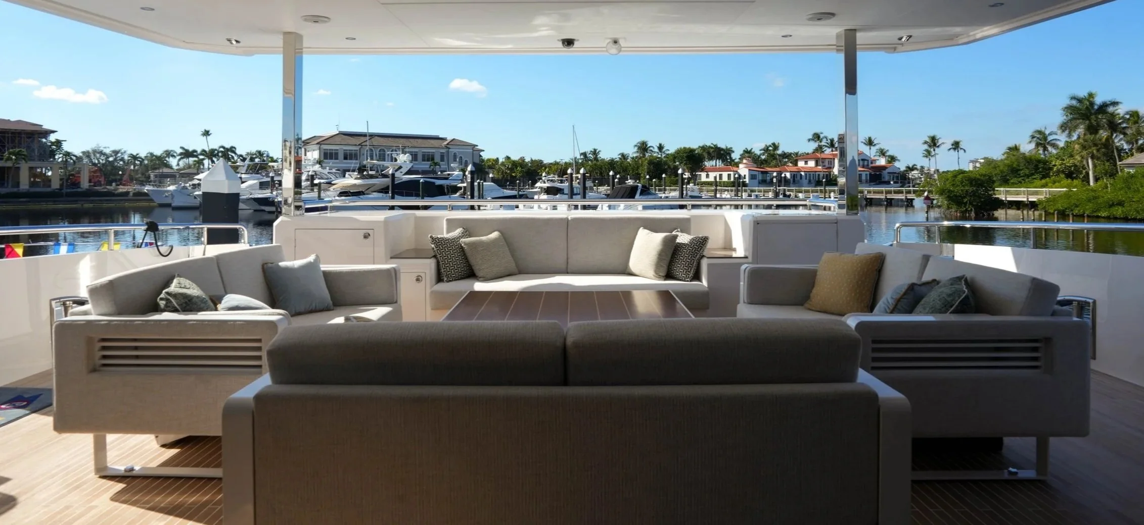Luxury yacht deck with white cushioned seating and pillows, overlooking a marina with moored boats, palm trees, and residential buildings under a clear blue sky.