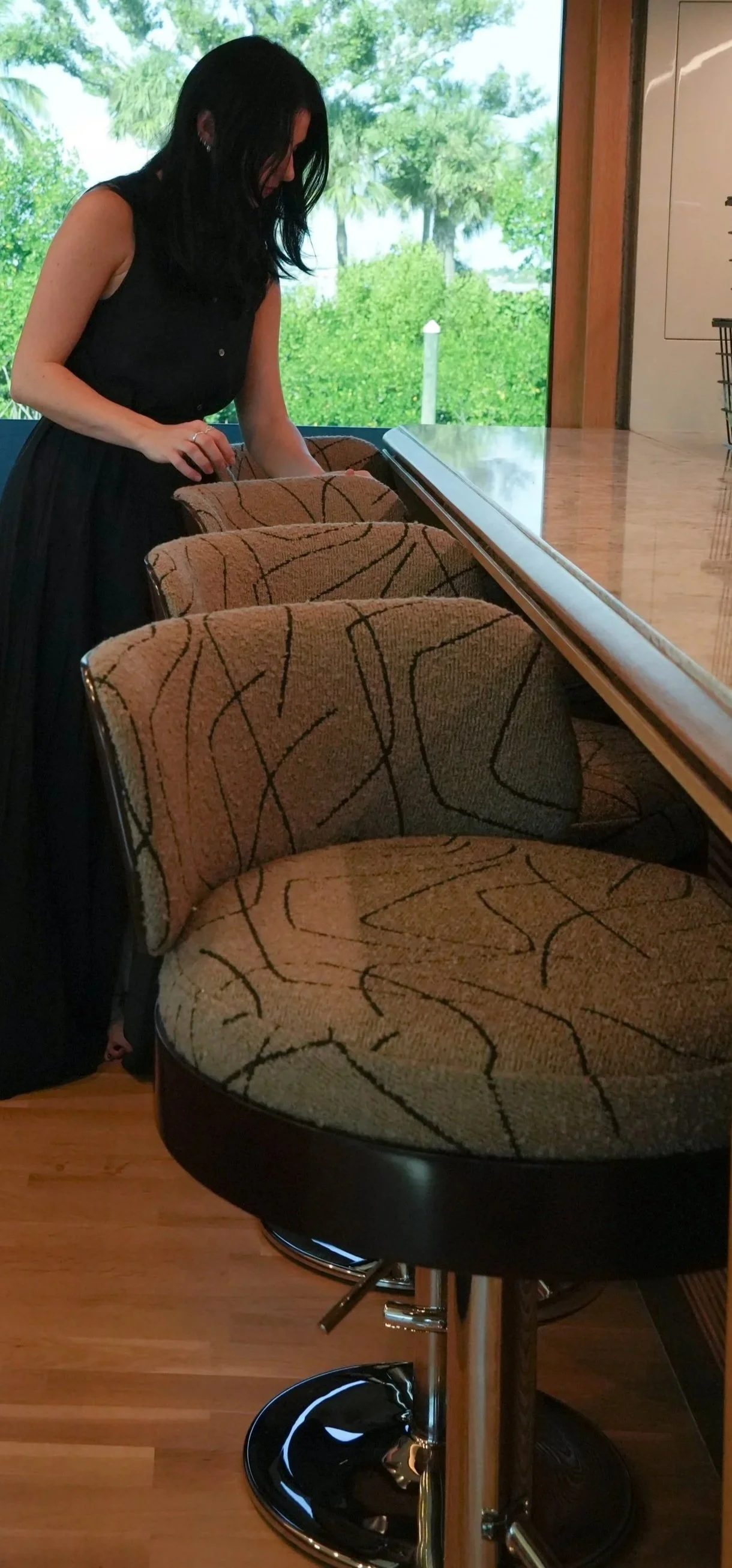 A woman with black hair, wearing a black sleeveless dress, leaning over a row of patterned barstools near a high counter, with a large window showing greenery outside.