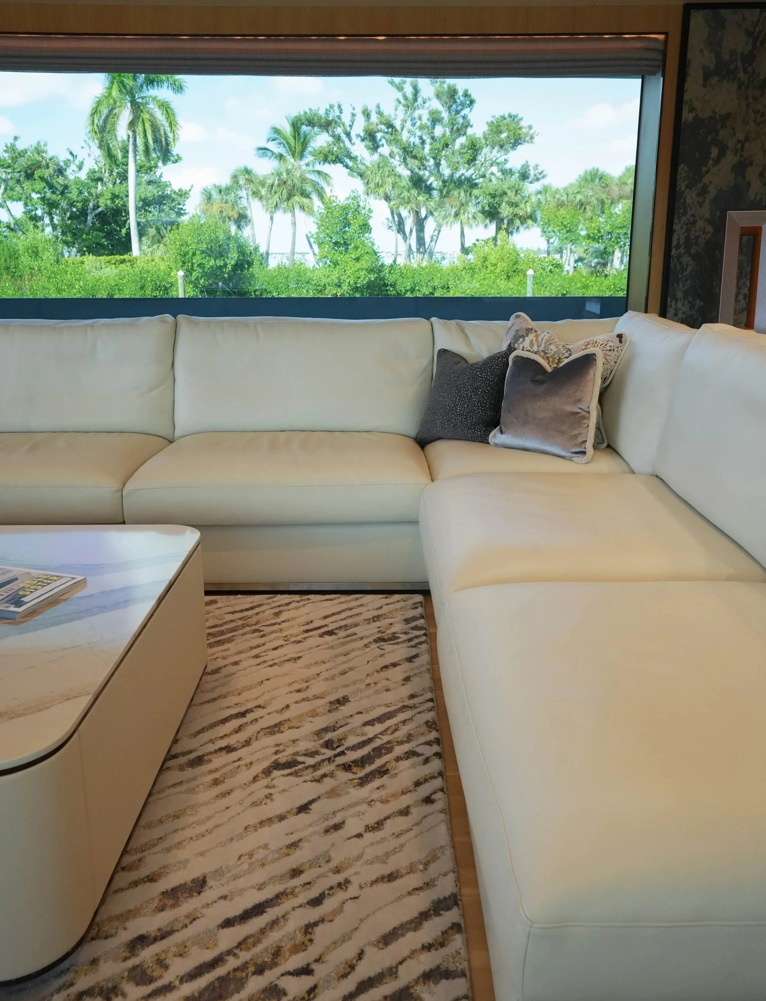 A living room with a large beige sectional sofa and decorative pillows, a marble coffee table, and a large window with a view of lush green trees and blue sky.