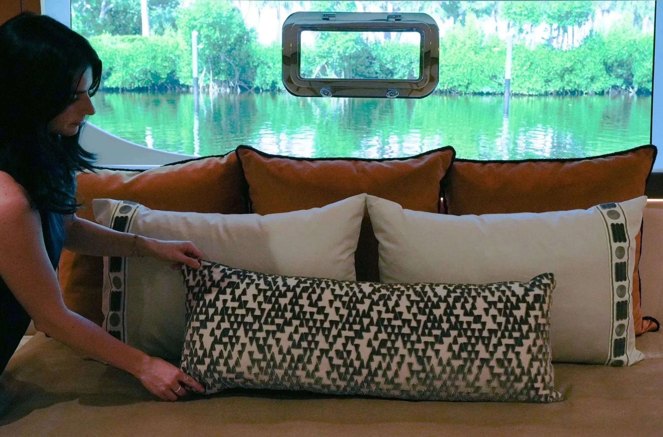 Woman adjusting decorative cushions on a bed or sofa with a window showing trees and water in the background.