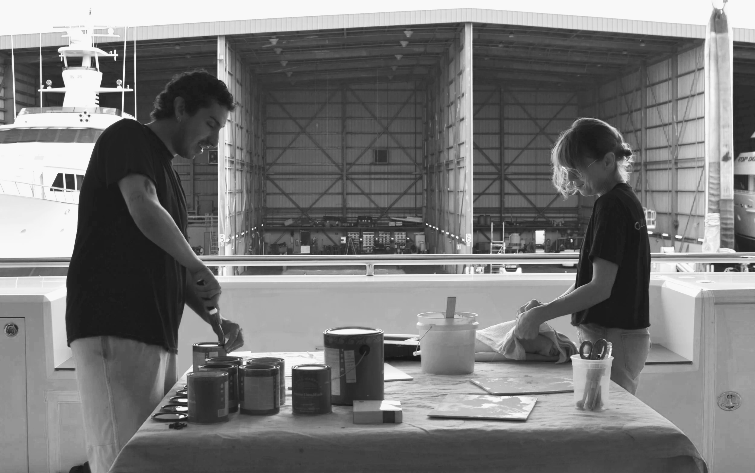 Two people, a man and a woman, standing behind a table inside a boat hangar, preparing or working with various cans and tools, with large ships visible in the background.