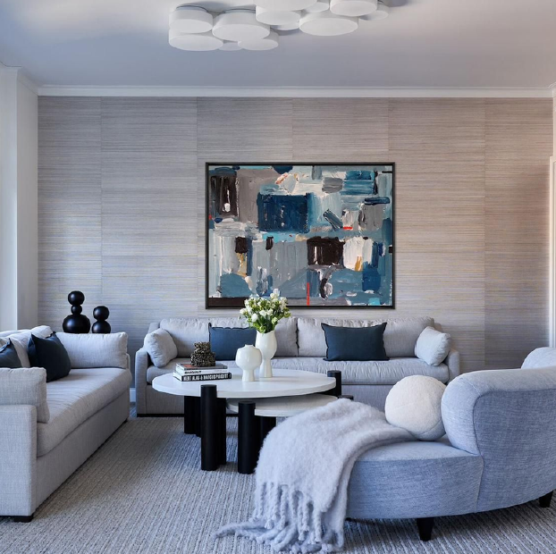 A modern living room with light gray sofas, a round white coffee table with decorative vases, navy and white pillows, and abstract art on the wall. There is a textured beige wall and a ceiling light fixture.