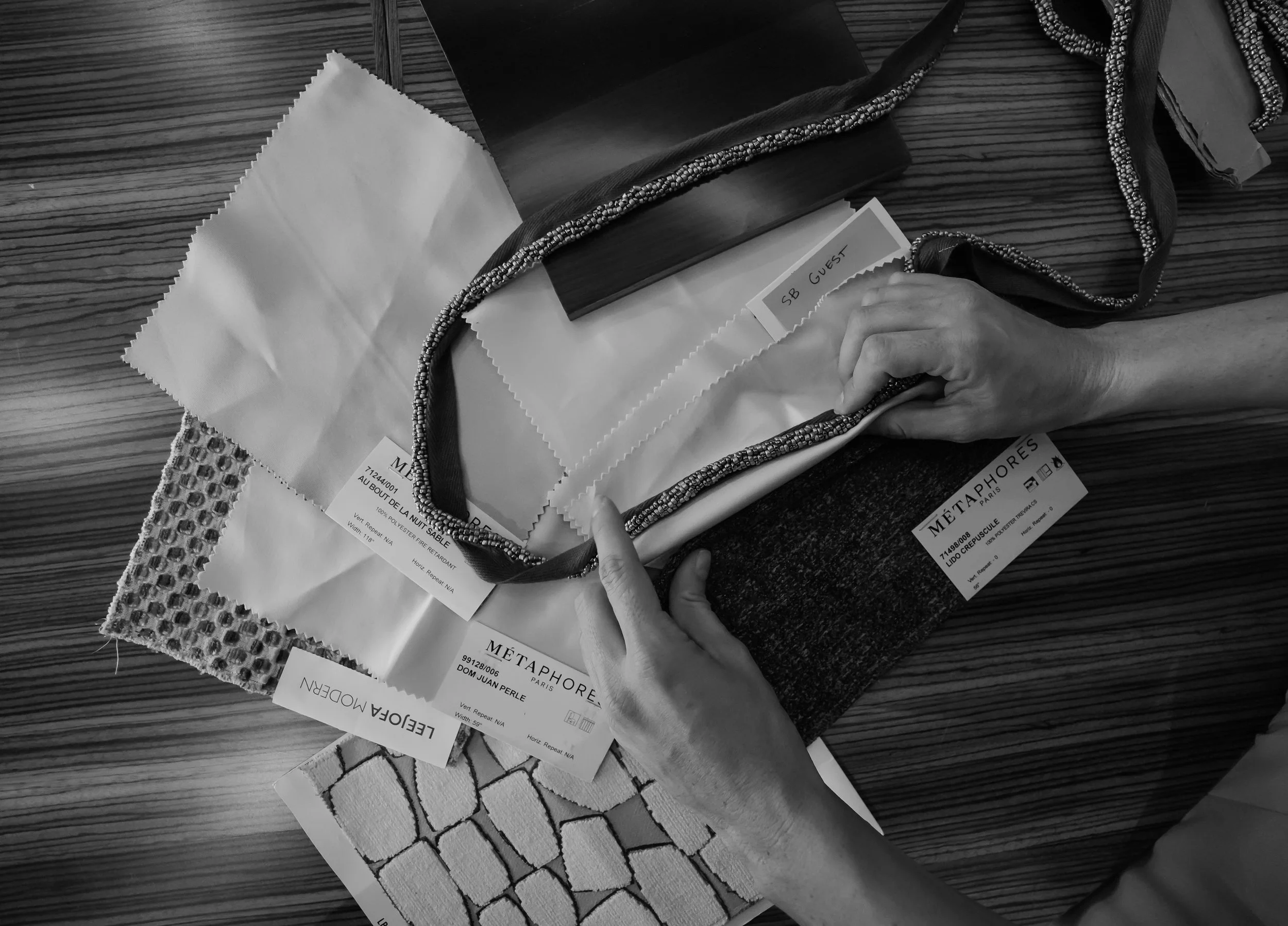 A person's hands holding a beaded necklace, with various fabric samples and labels on a wooden table.
