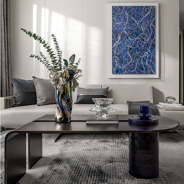 A modern living room with a white sofa, gray pillows, a black coffee table with a colorful vase of greenery and dried flowers, a blue glass cup on a dark coaster, a glass bowl, and two books. There is an abstract blue painting on the wall.