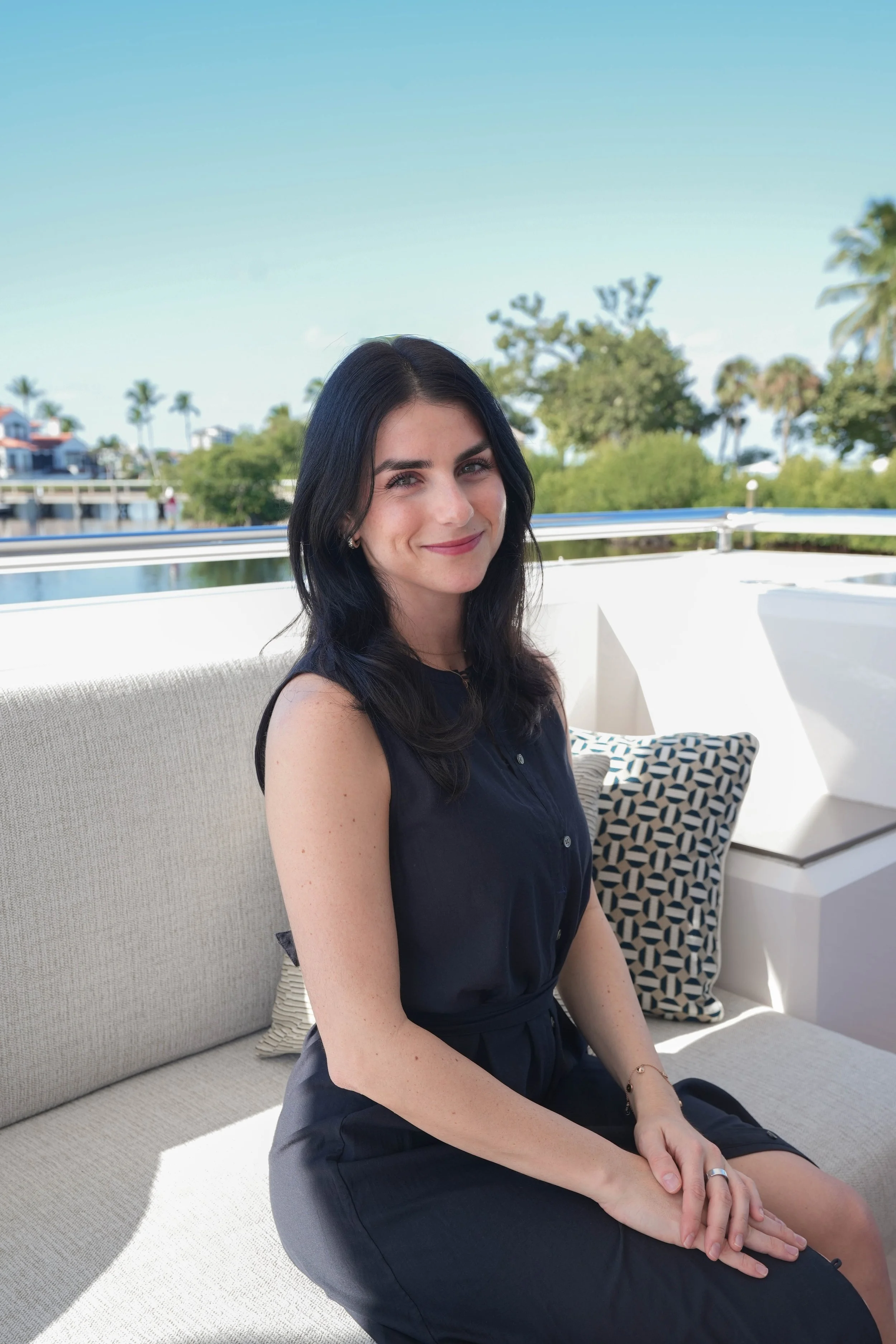 A woman with black hair sitting on a beige outdoor couch, smiling, with a river, trees, and palm trees in the background on a sunny day.