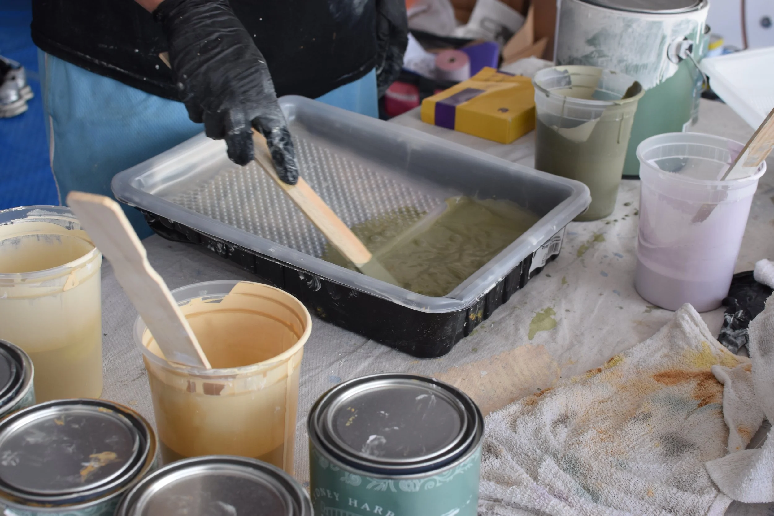 Person stirring liquid in a plastic tray at a workshop with various containers of paint and tools on the table.
