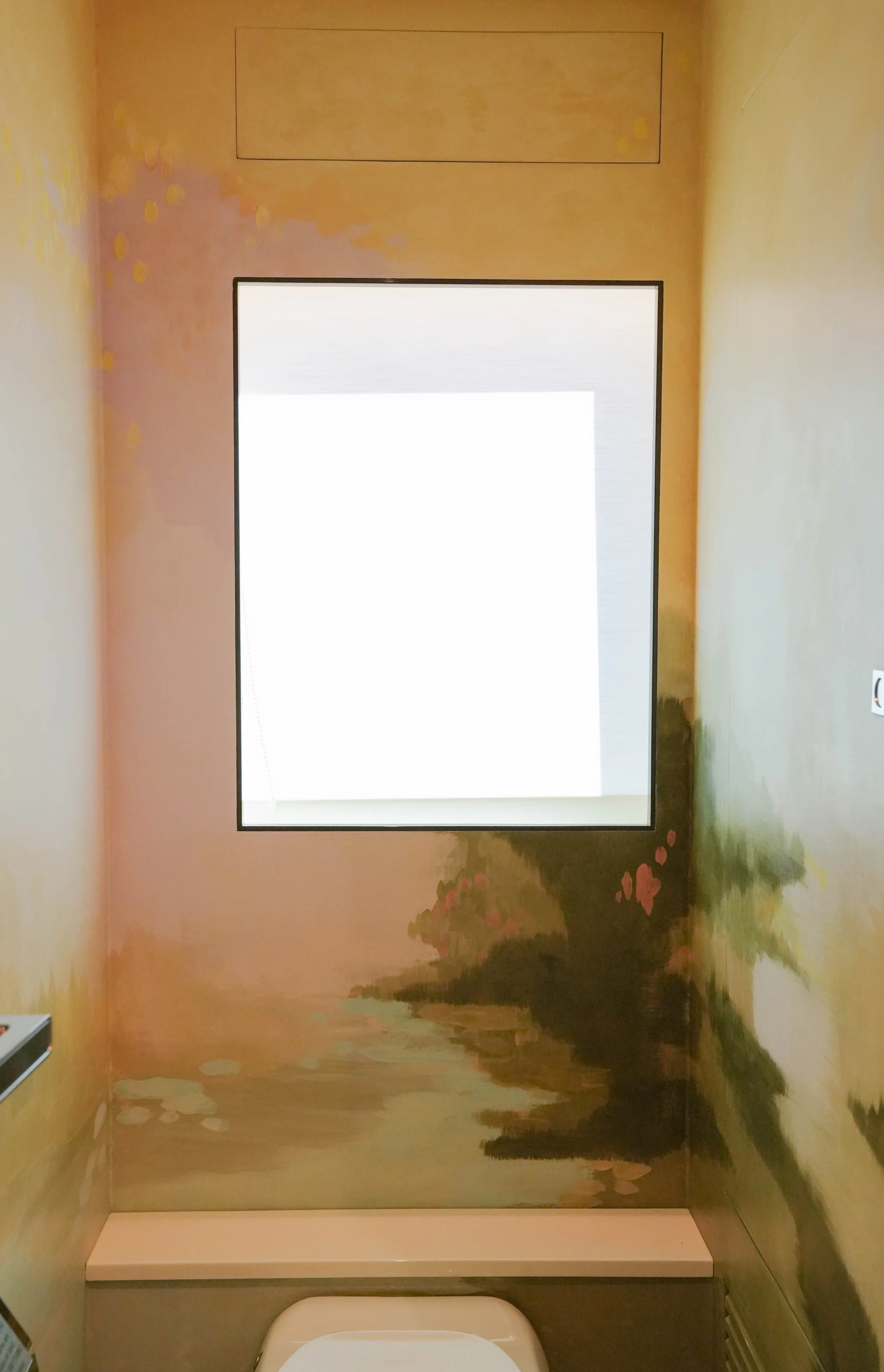 Interior view of a bathroom with a wall painting of a landscape, a window, and a toilet.