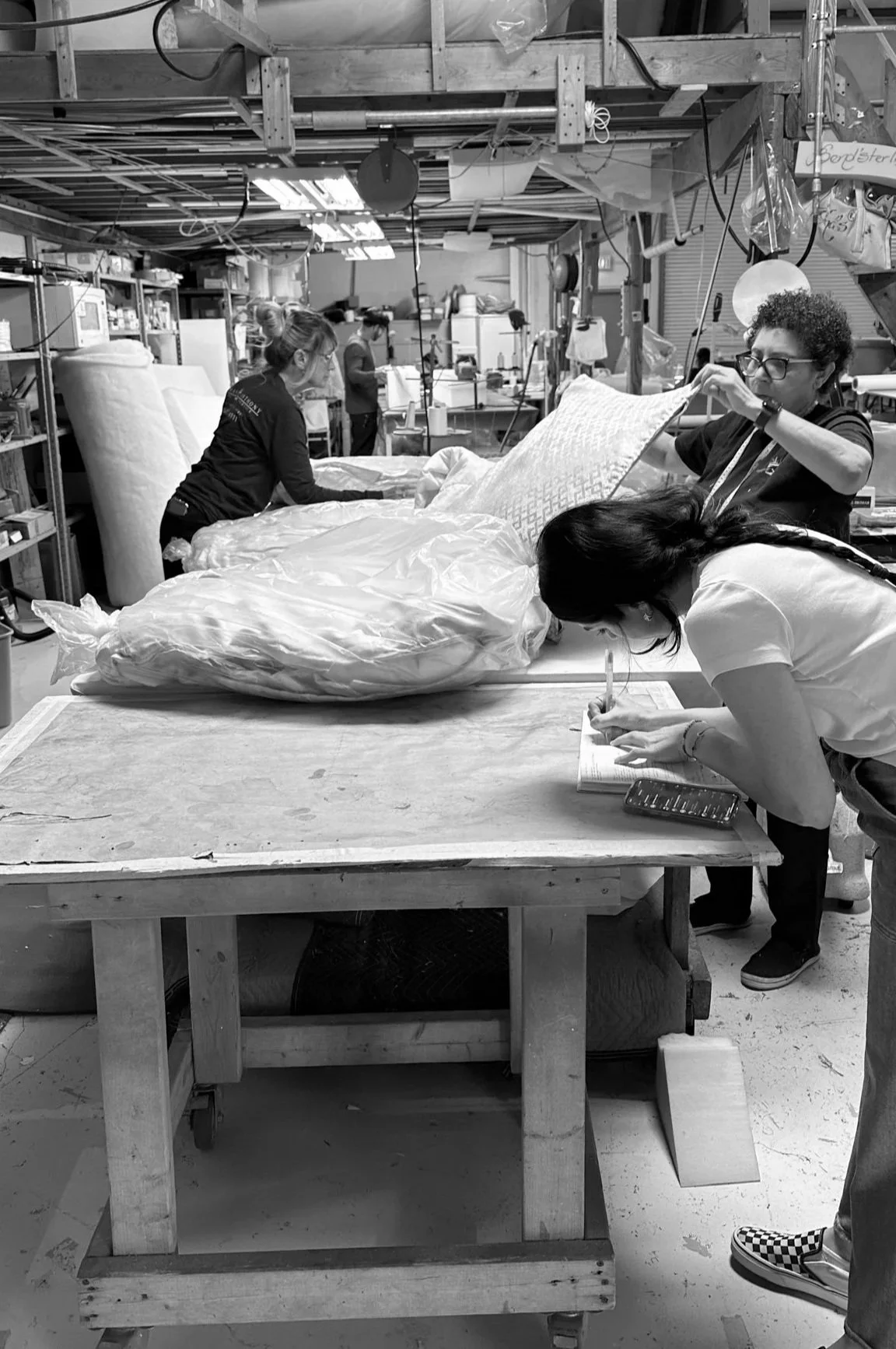 People working in a workshop, folding a large fabric object, surrounded by shelves and equipment.