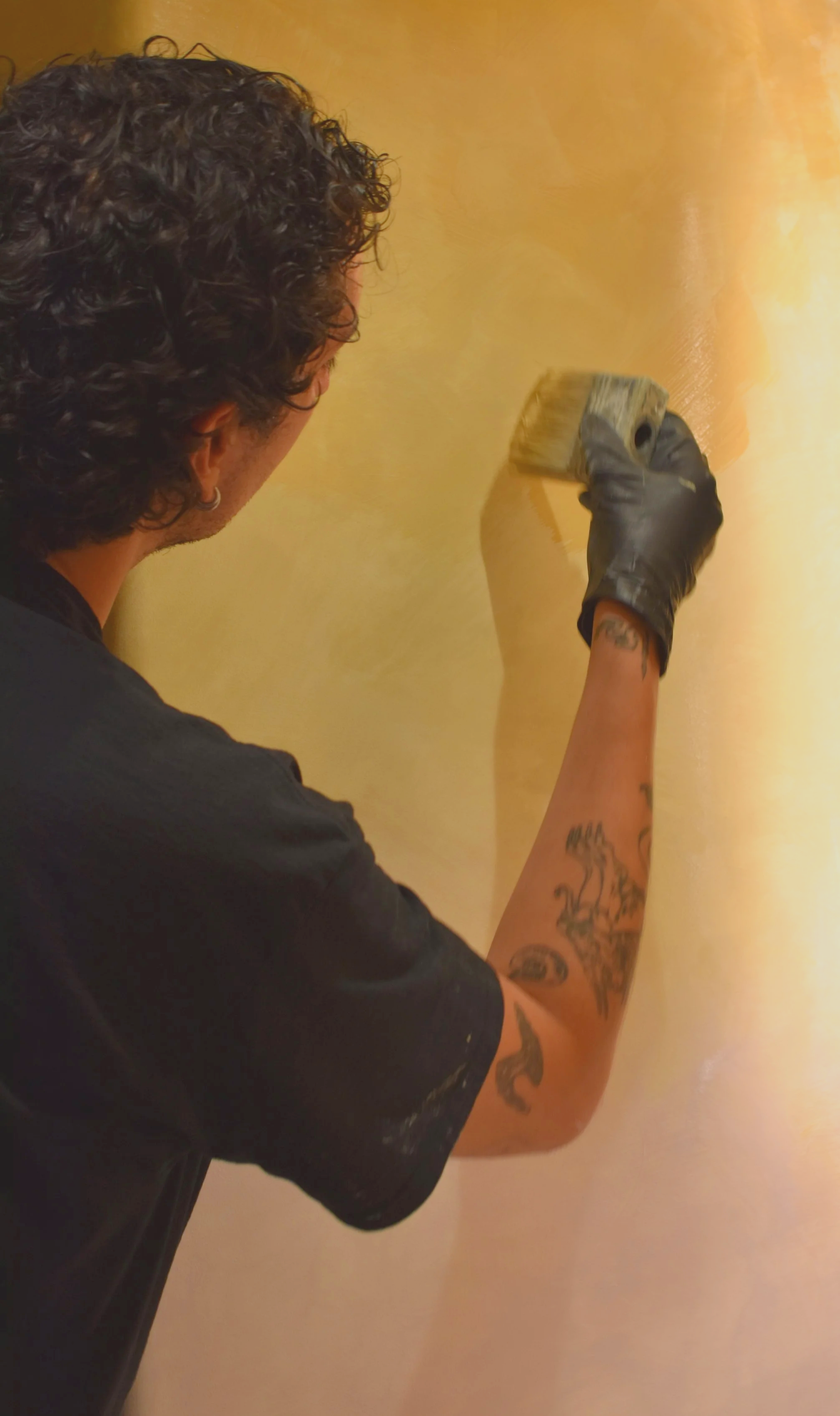 Person with curly hair and tattoos painting a wall with a paintbrush.