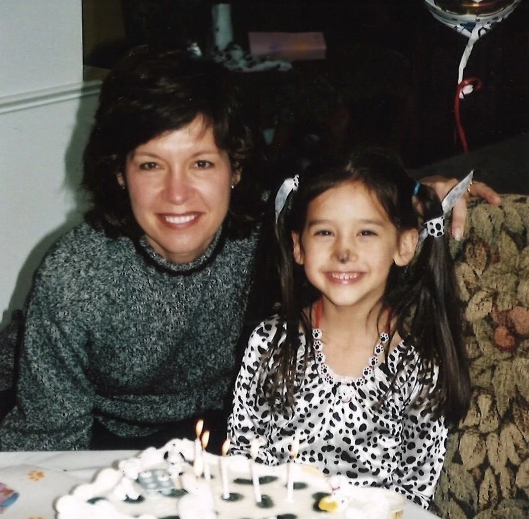 A woman and a young girl smiling at a birthday celebration, with a birthday cake with lit candles in the foreground.