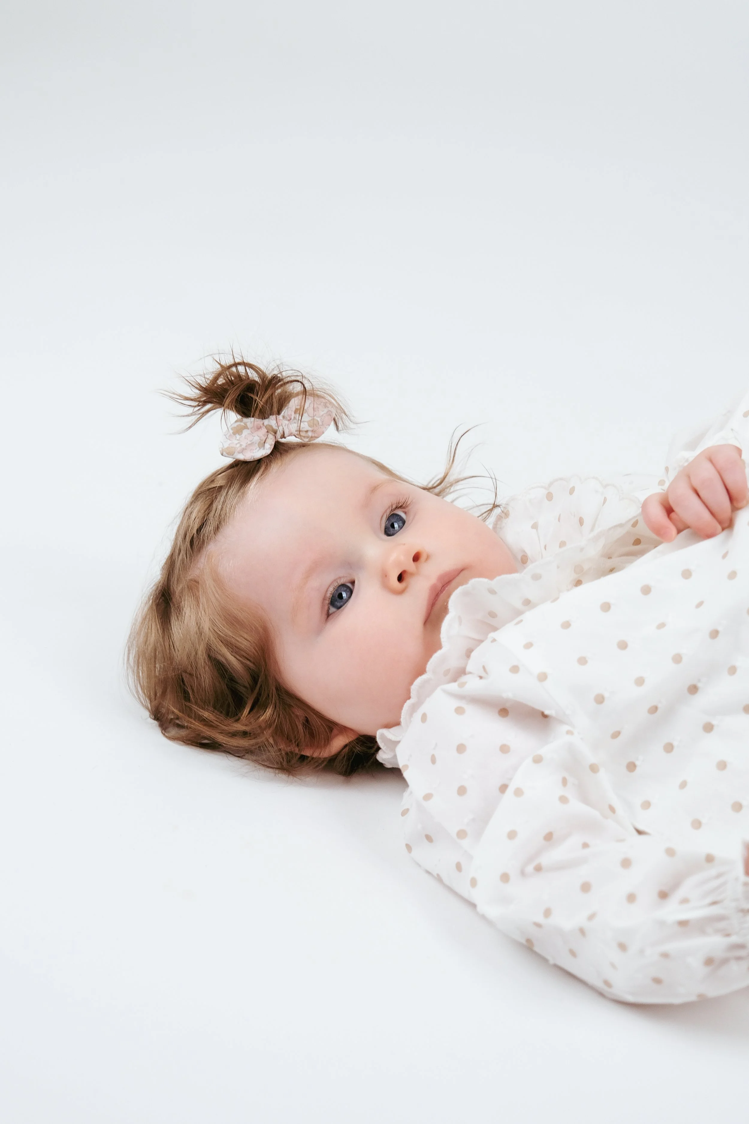 A young girl with light brown curly hair, blue eyes, and a pink hair scrunchie lying on a white surface, wearing a white polka-dot dress