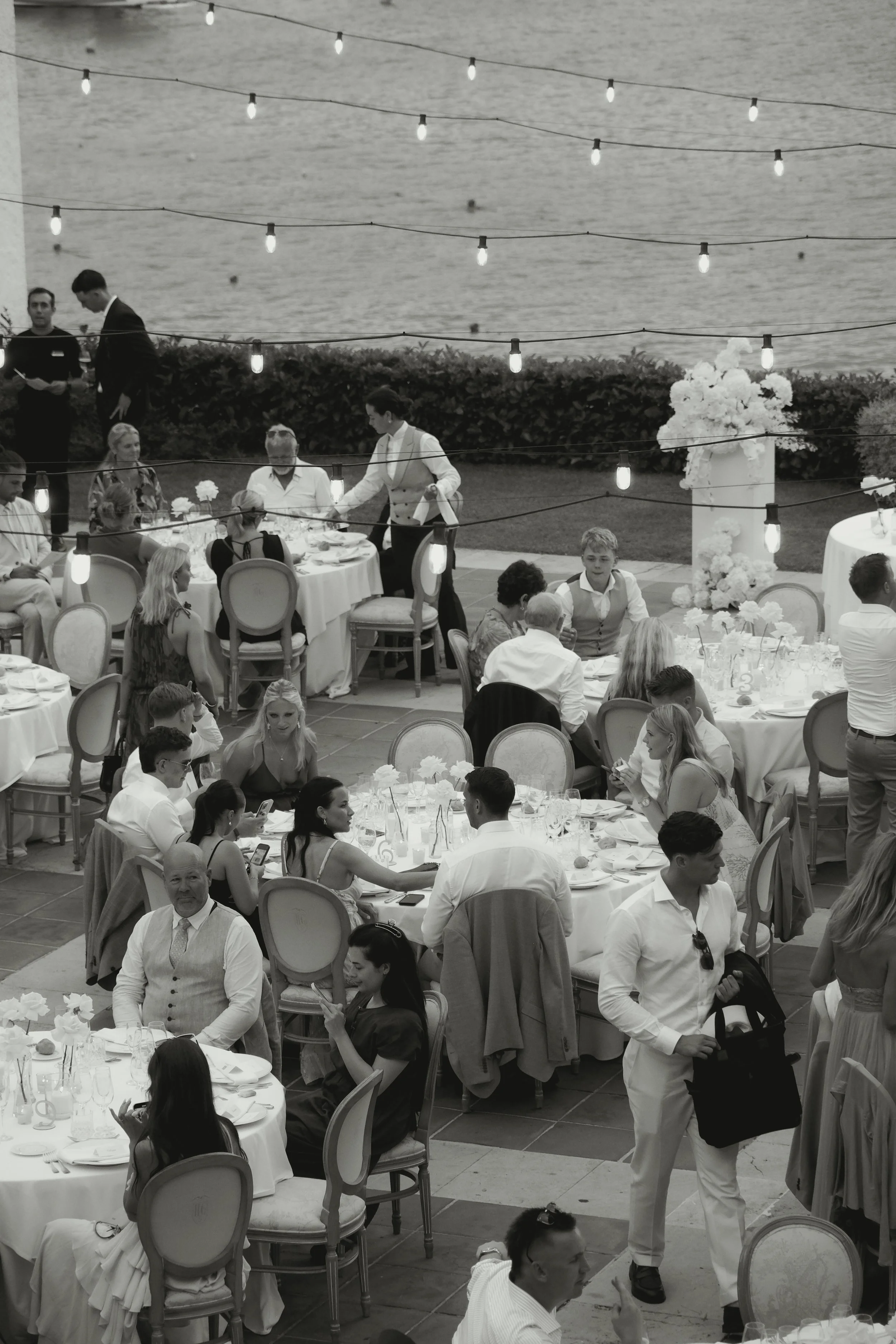 A black and white photo of a lively outdoor dinner event by the water, with decorated tables, guests seated and mingling, and string lights overhead.