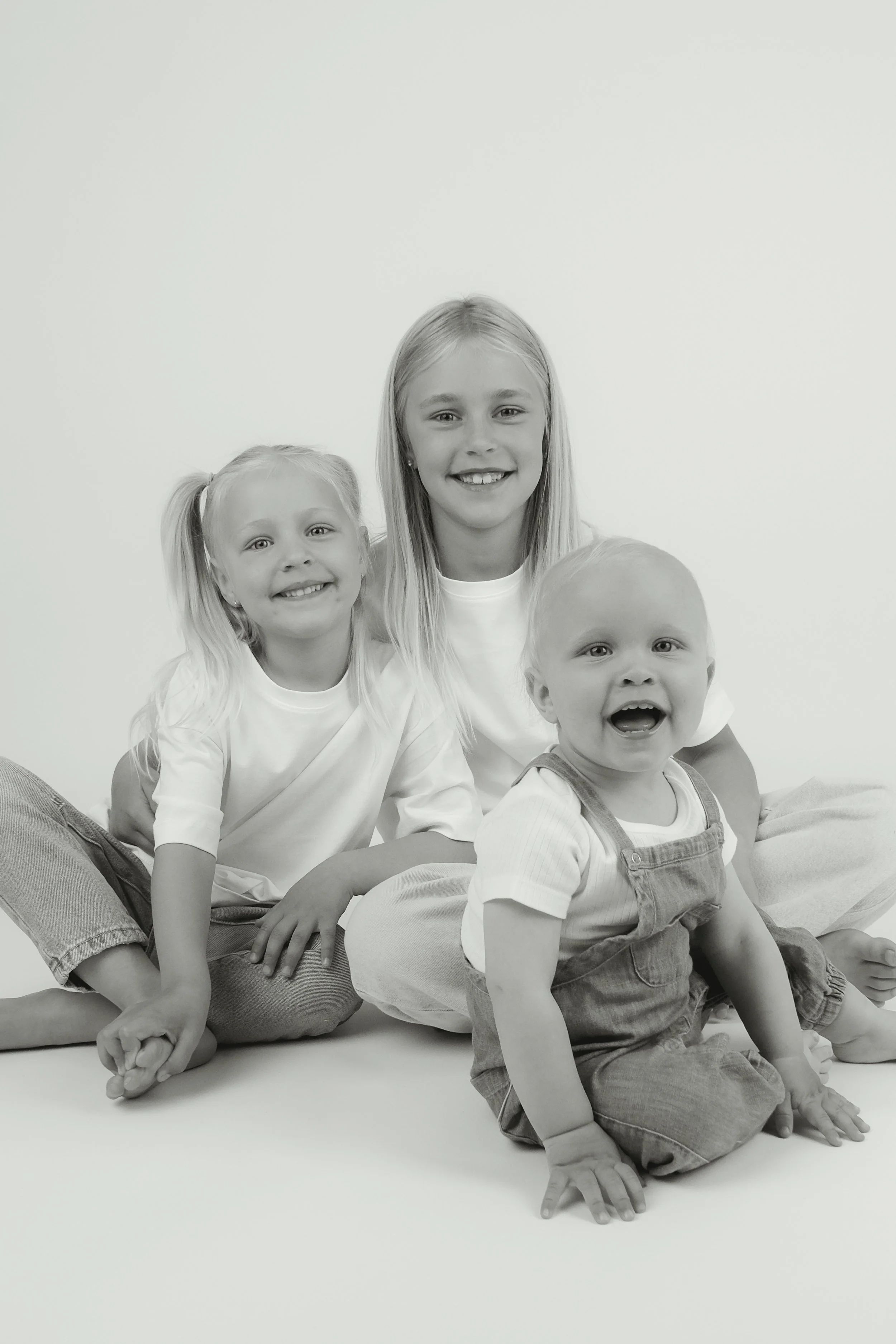 Black and white photo of three smiling children sitting on the floor against a plain background. Two older girls and a younger boy.