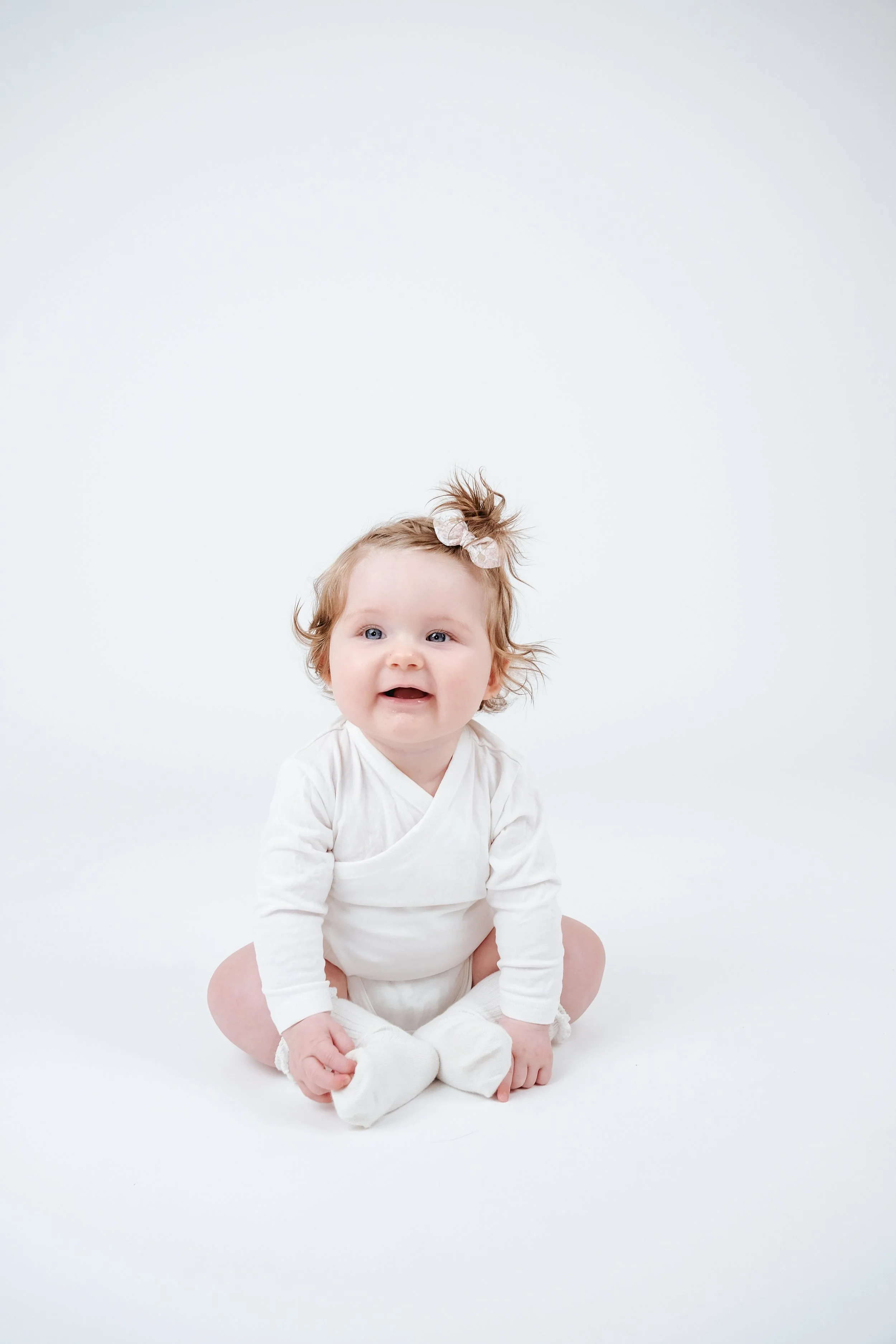 A smiling baby girl sitting on a white background, dressed in a white outfit with a bow in her hair.
