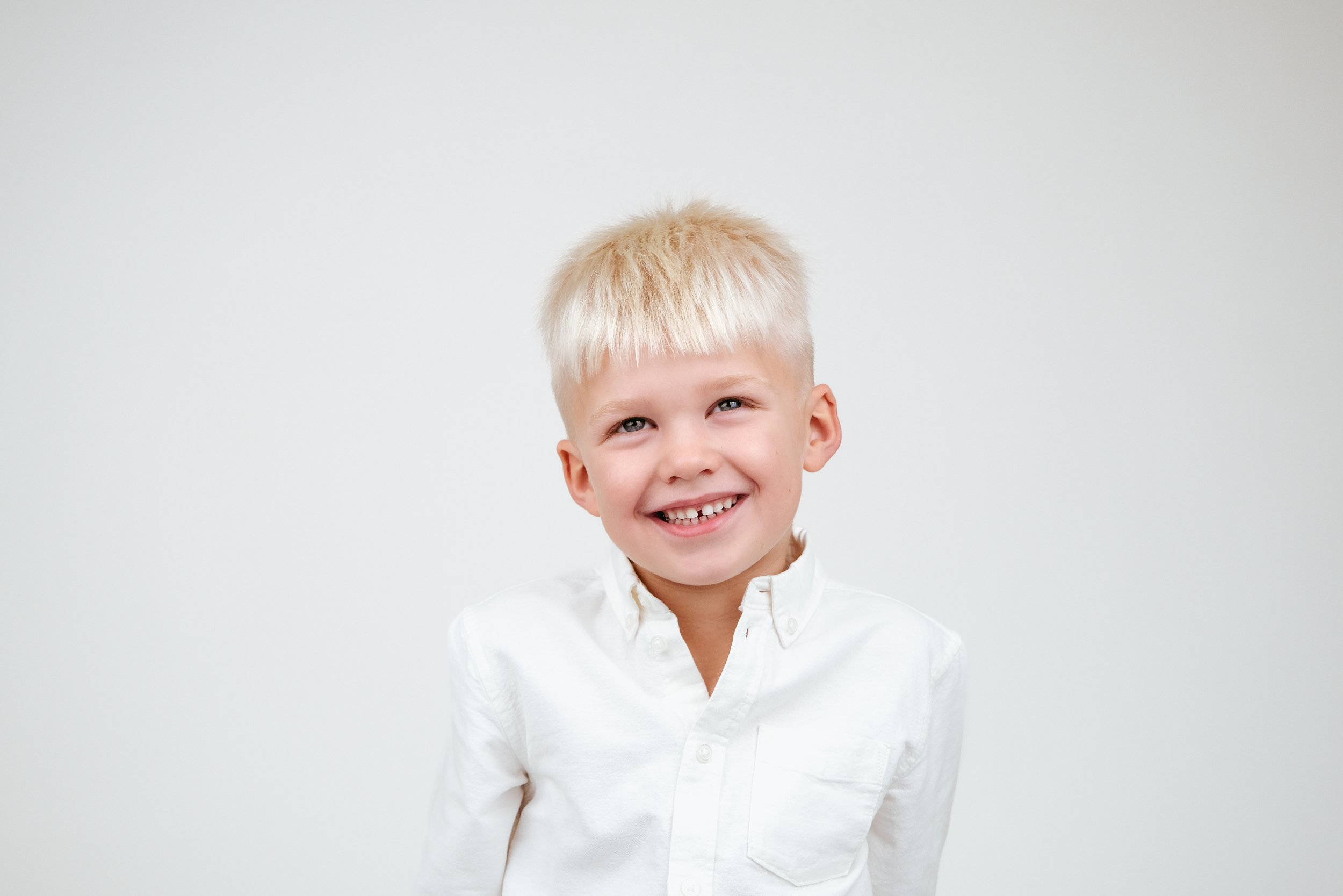 Young boy with blonde hair smiling, wearing a white button-up shirt, against a plain white background.