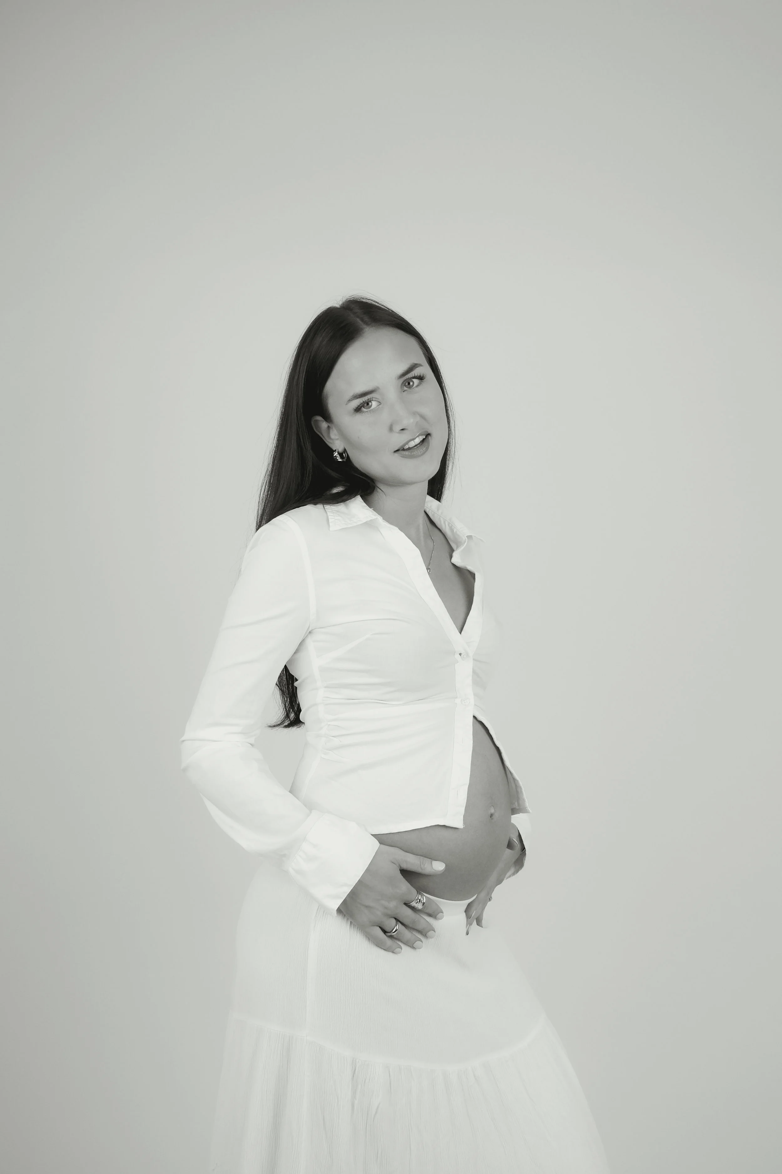 A pregnant woman wearing a white button-up shirt and a long skirt, posing against a plain background, looking at the camera with a slight smile.