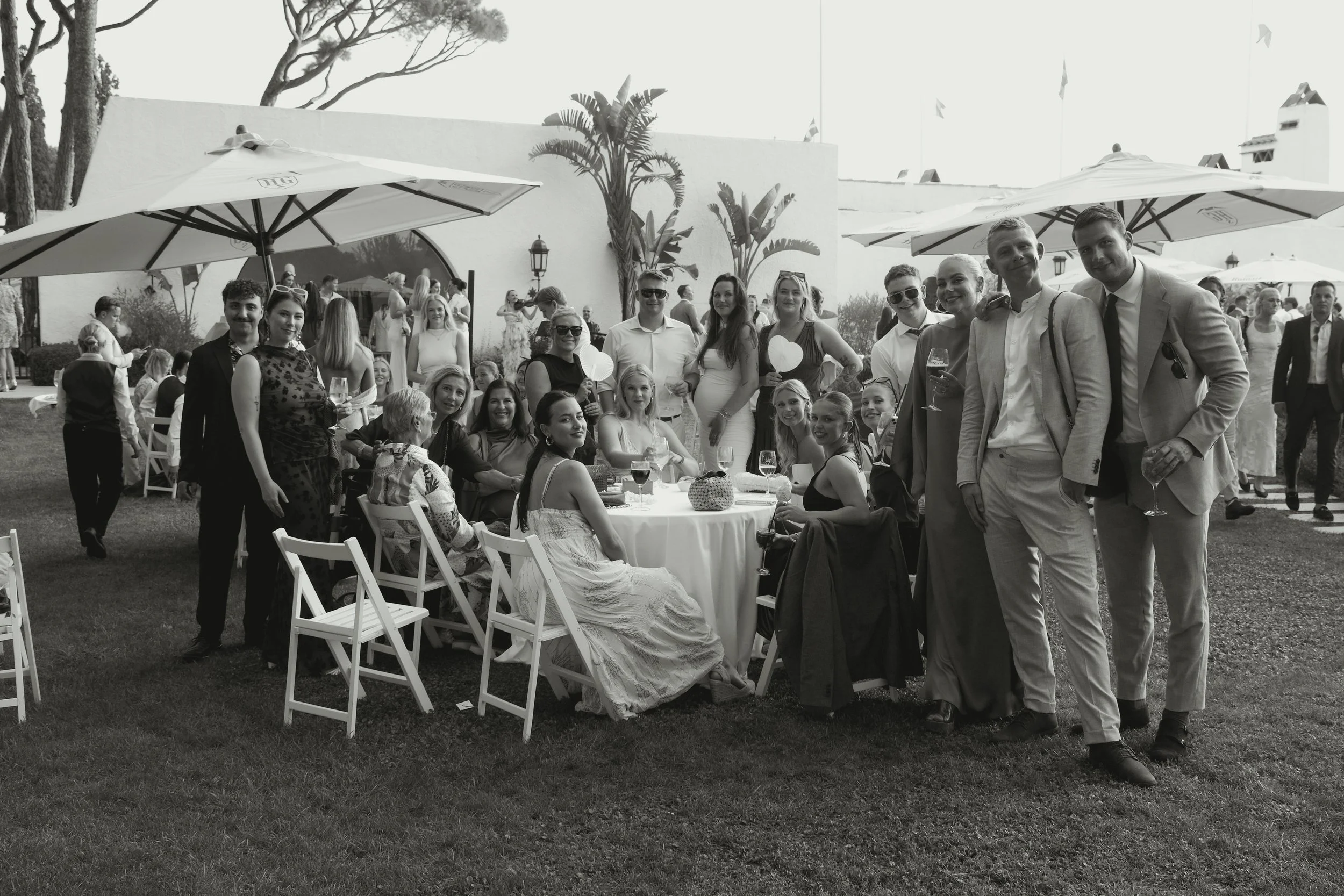 Group of people gathered at a wedding reception outdoors, sitting and standing around a table with drinks, with umbrellas and palm trees in the background.