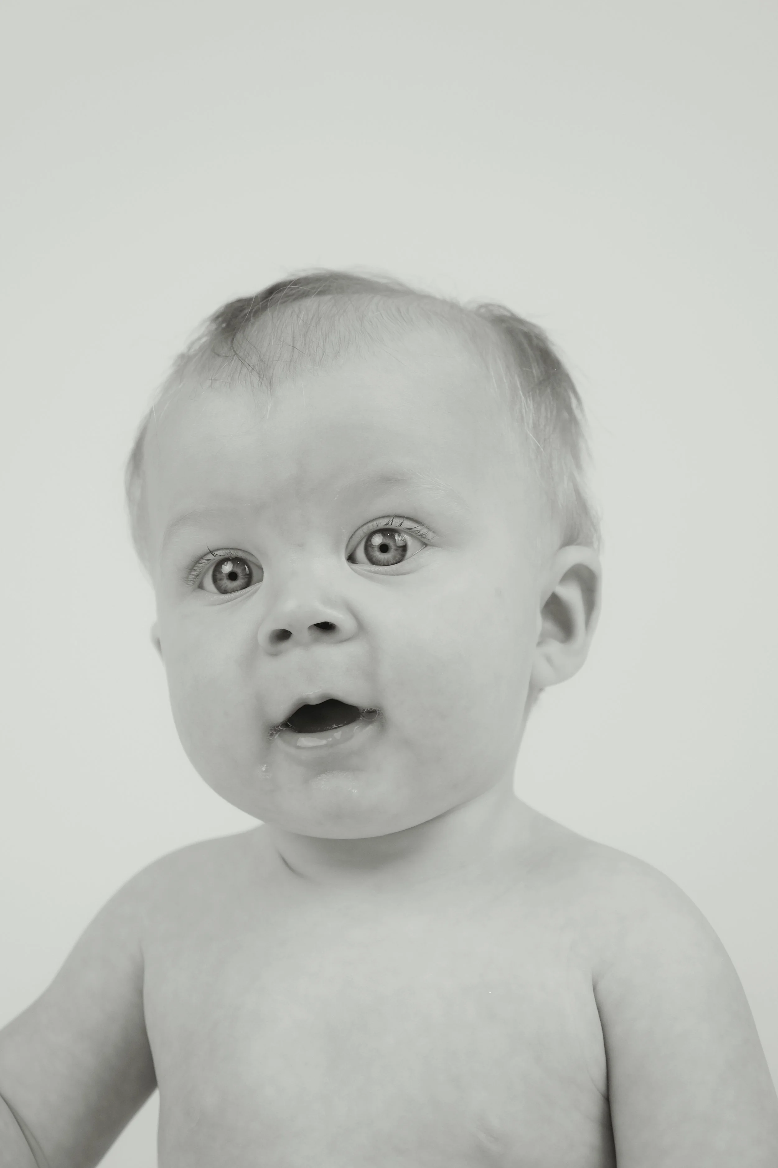 Close-up black-and-white photo of a baby with surprised expression, wide eyes, and open mouth, without clothing.