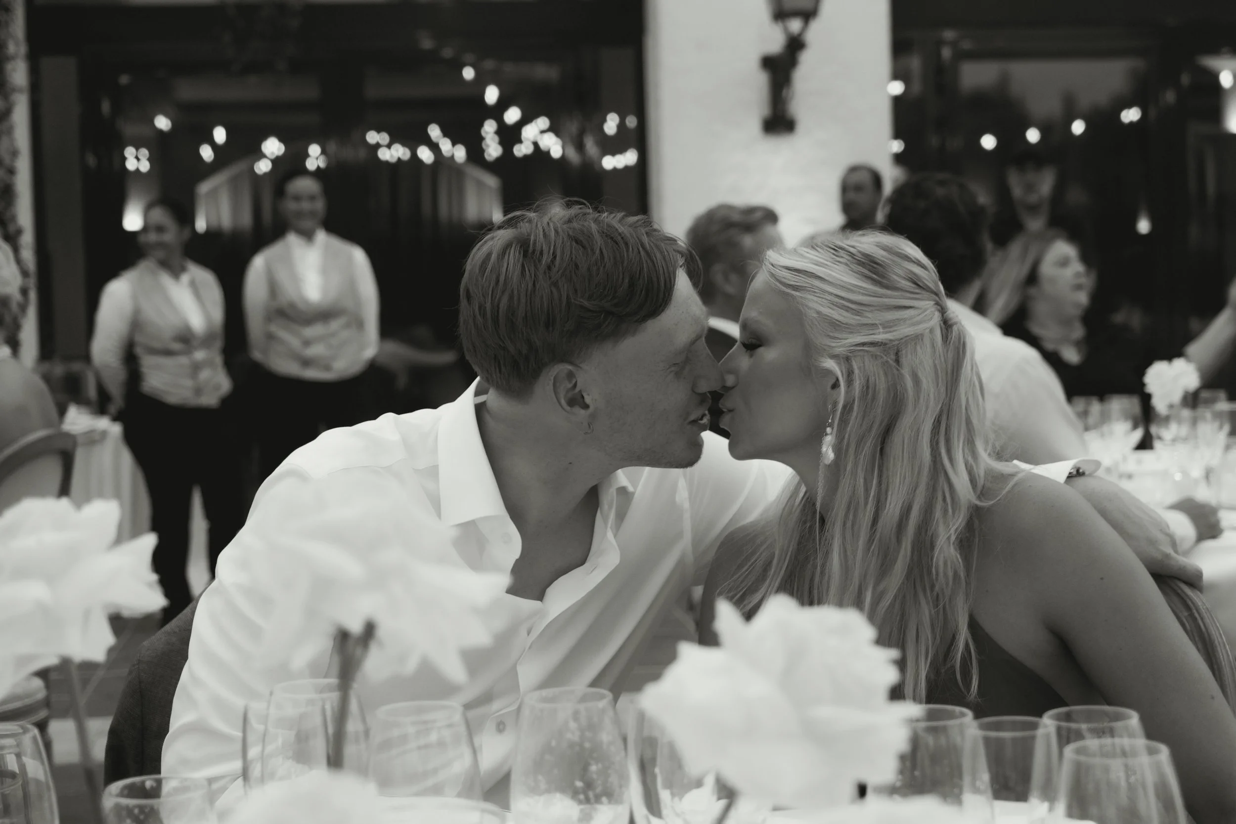 A black and white photo of a couple about to kiss at a wedding reception, with guests and waitstaff in the background.