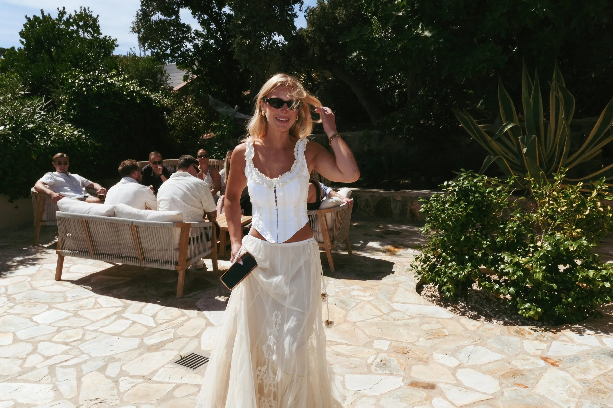 A smiling woman in sunglasses, a white sleeveless top, and a long skirt walking outdoors on a sunny day, holding a smartphone. In the background, a group of people is sitting on a patio with lush greenery around.