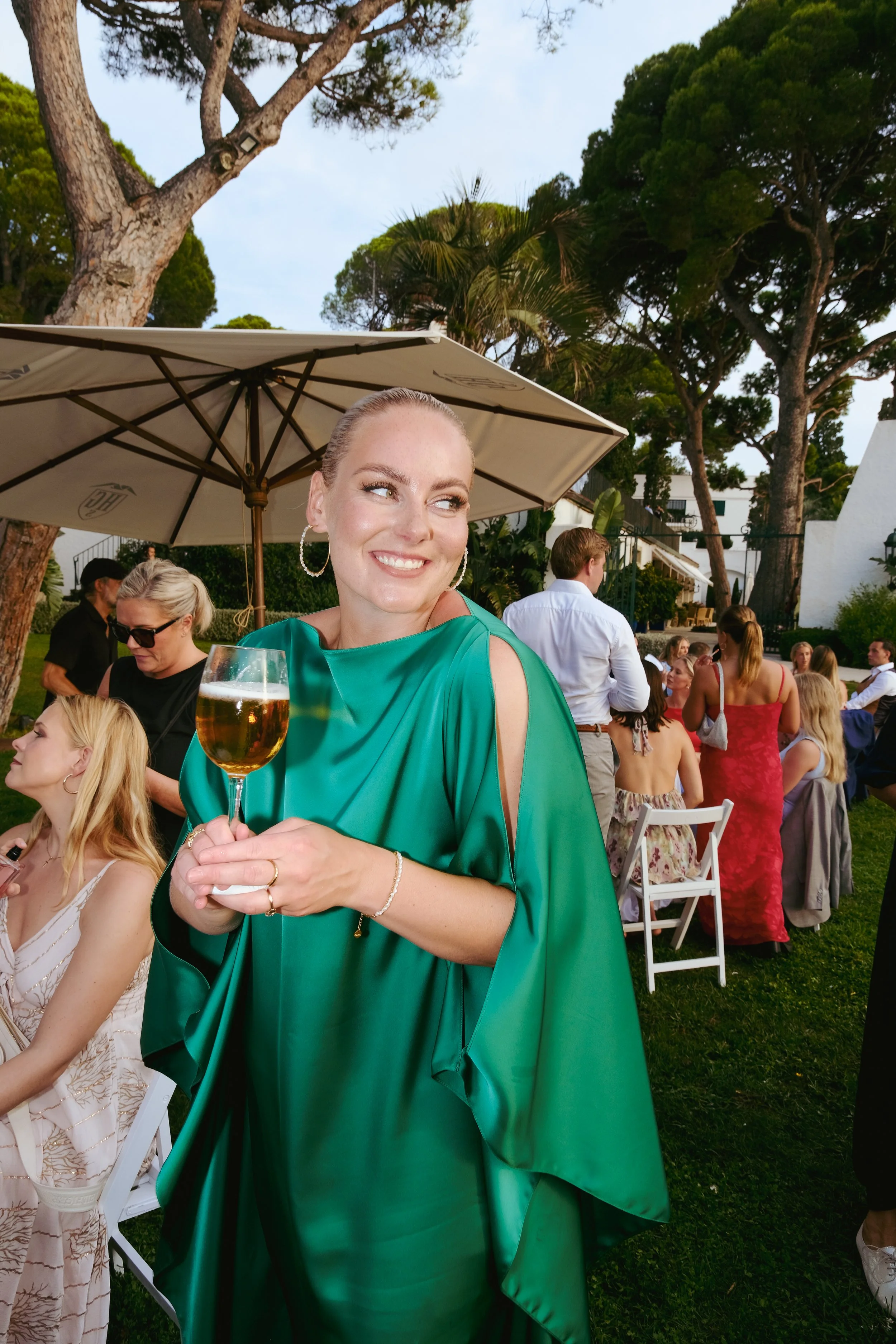 A woman in a green dress holding a glass of wine at an outdoor gathering with other guests and large trees in the background.