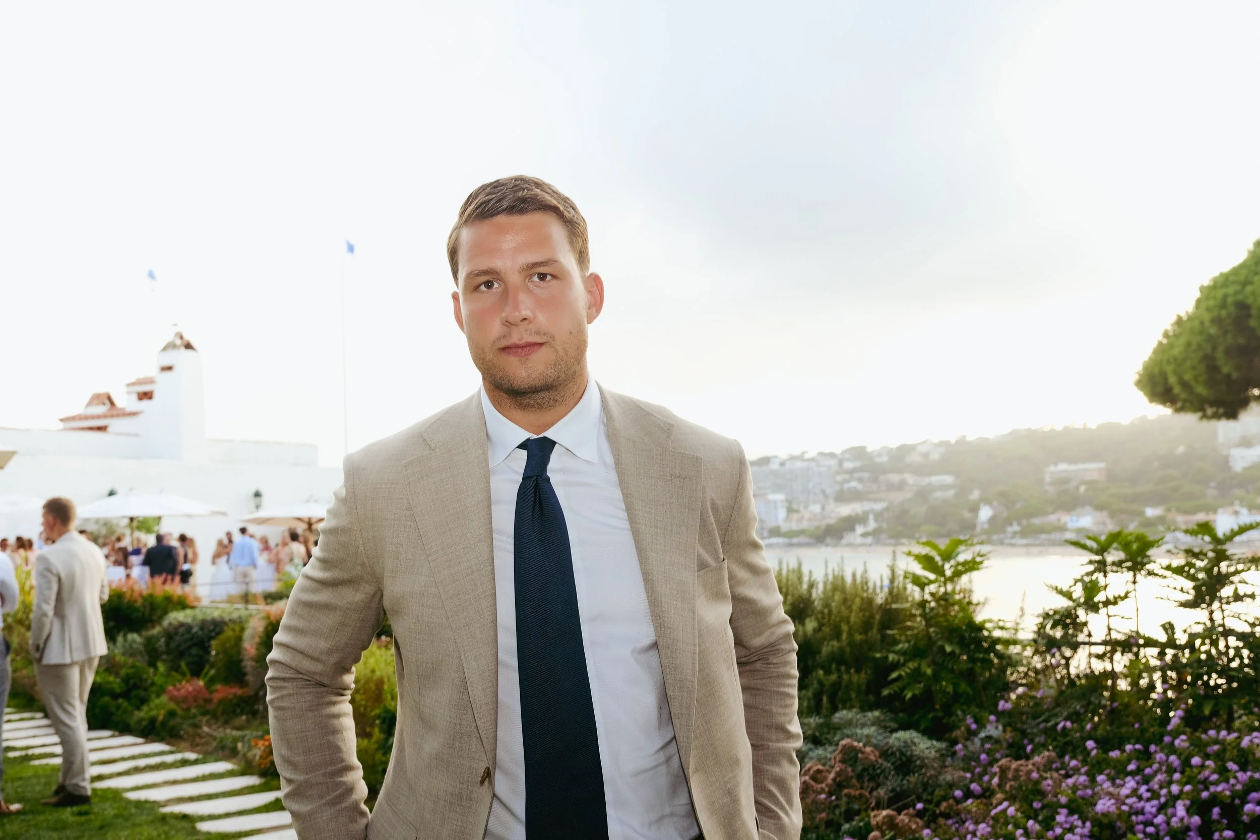 A man in a beige suit and blue tie standing outdoors at a social event, with a garden, water, hillside, and white building in the background.