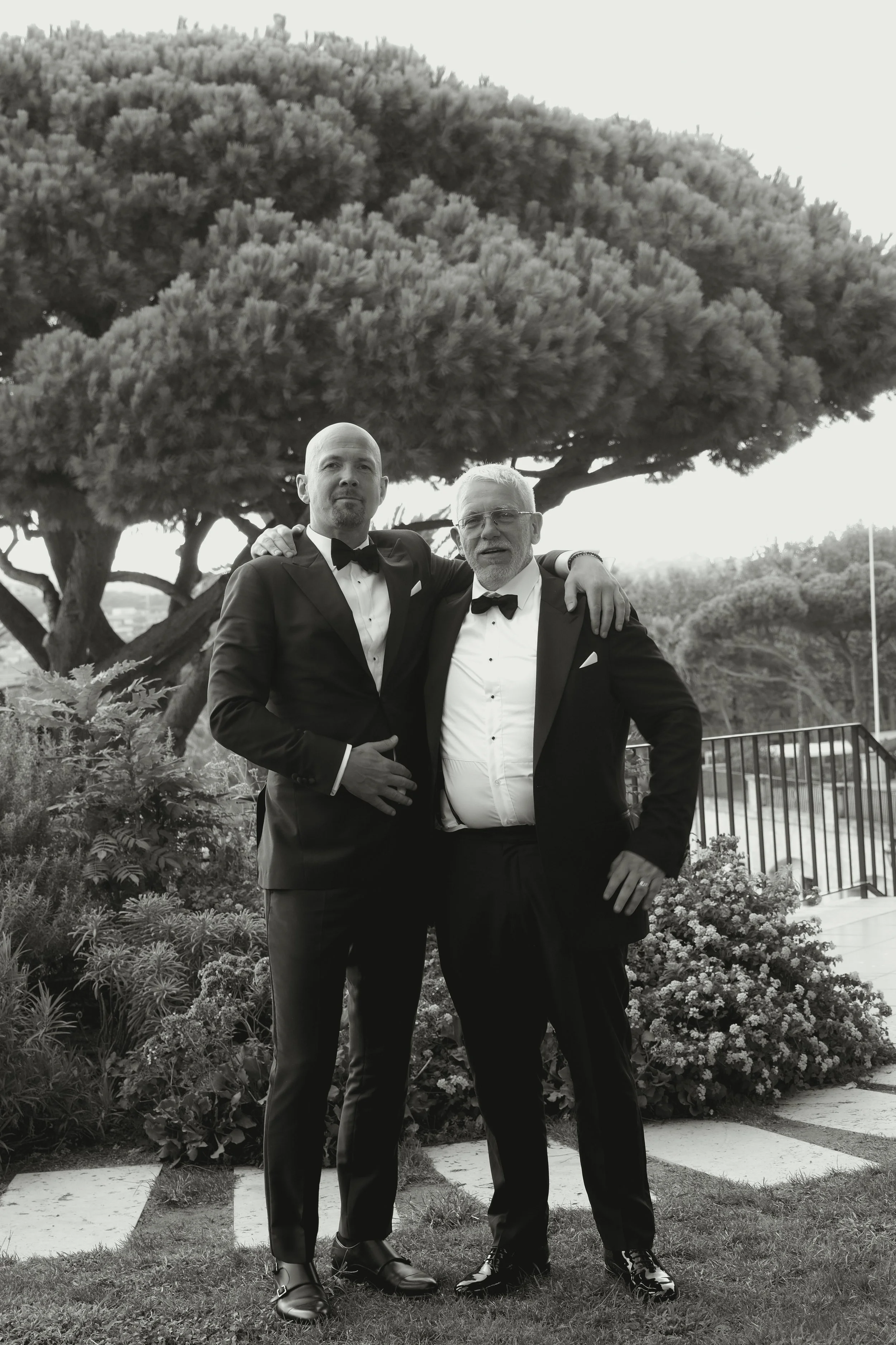 Two men in tuxedos with bow ties standing outdoors, posing with their arms around each other, in front of a large tree and garden plants.