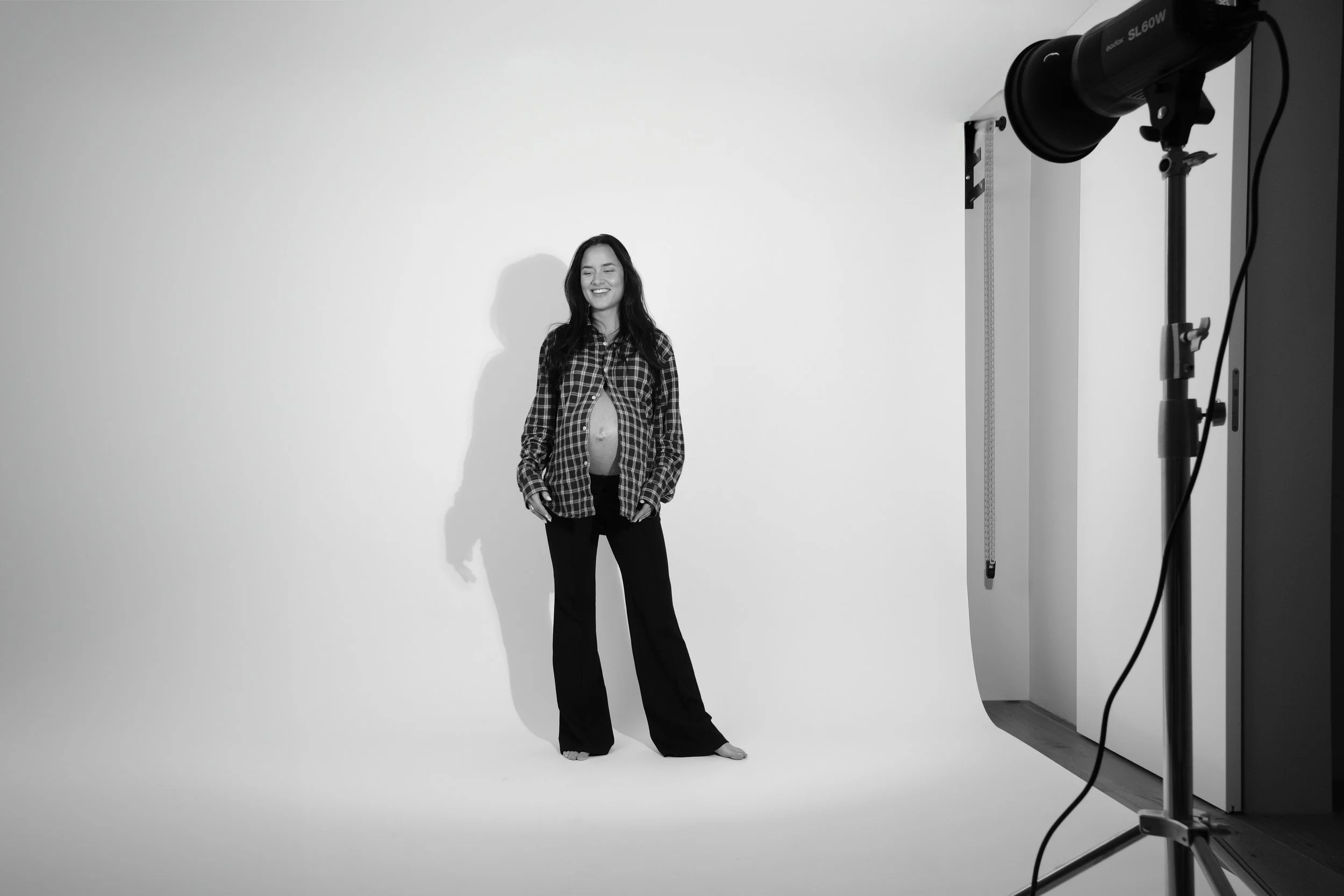 A pregnant woman smiling in a photography studio, standing in front of a plain white background, with photography equipment visible on the right side.