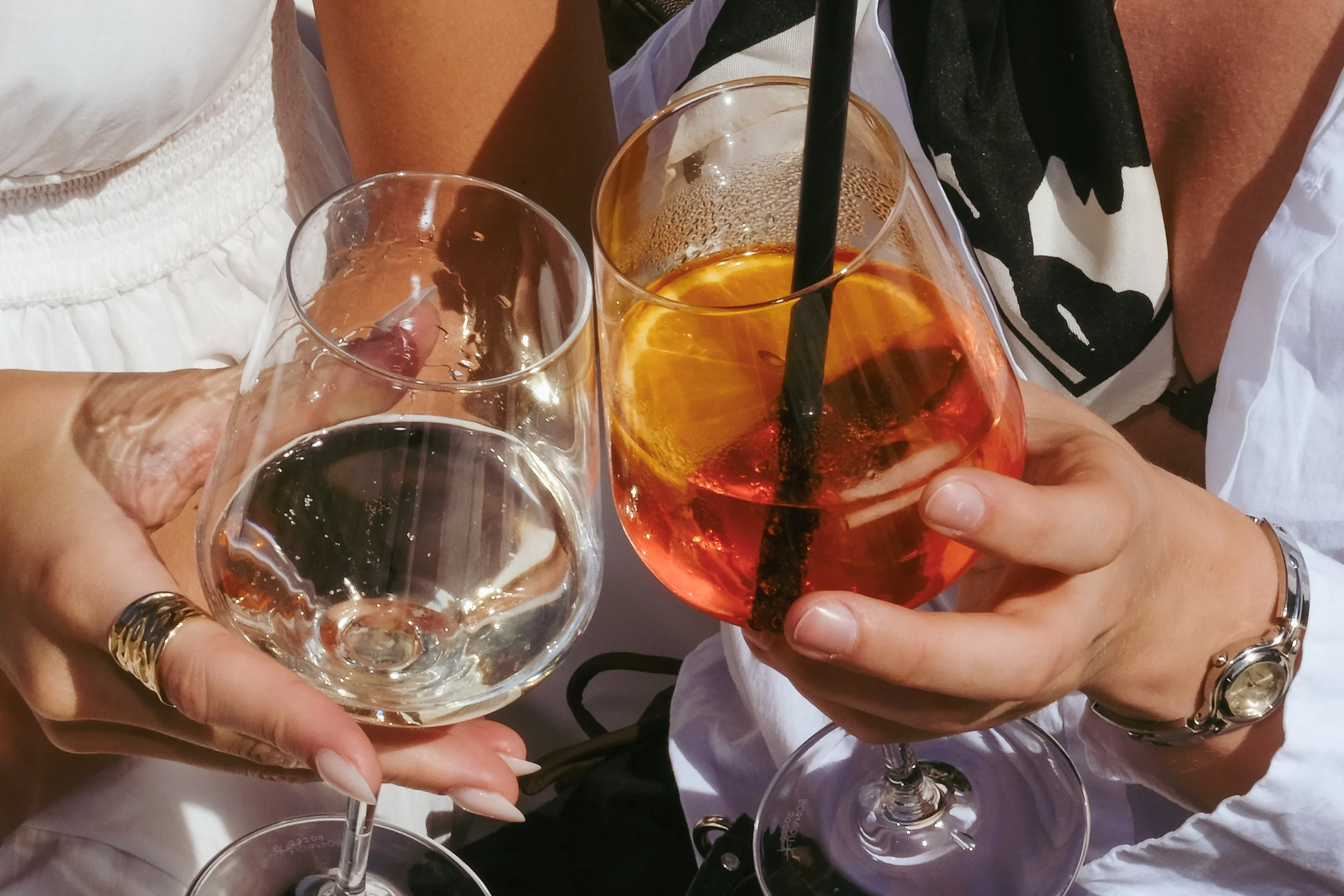 Two people holding glasses, one with a clear drink and the other with an orange-colored cocktail with a straw and lemon slice.