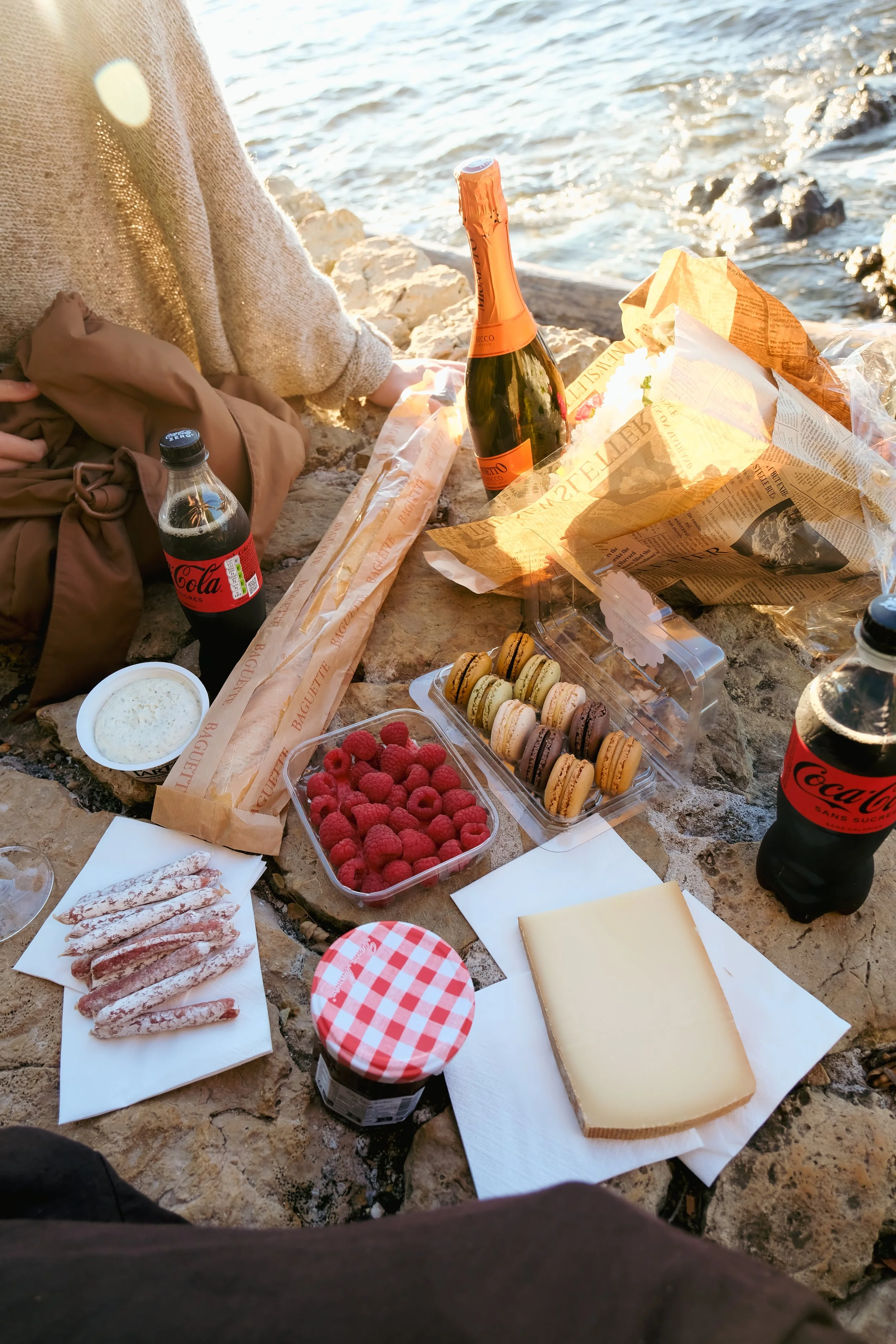 A seaside picnic setup with cheese, raspberries, macarons, snacks, Coca-Cola bottles, and a bottle of sparkling wine or champagne on a rocky surface near water.