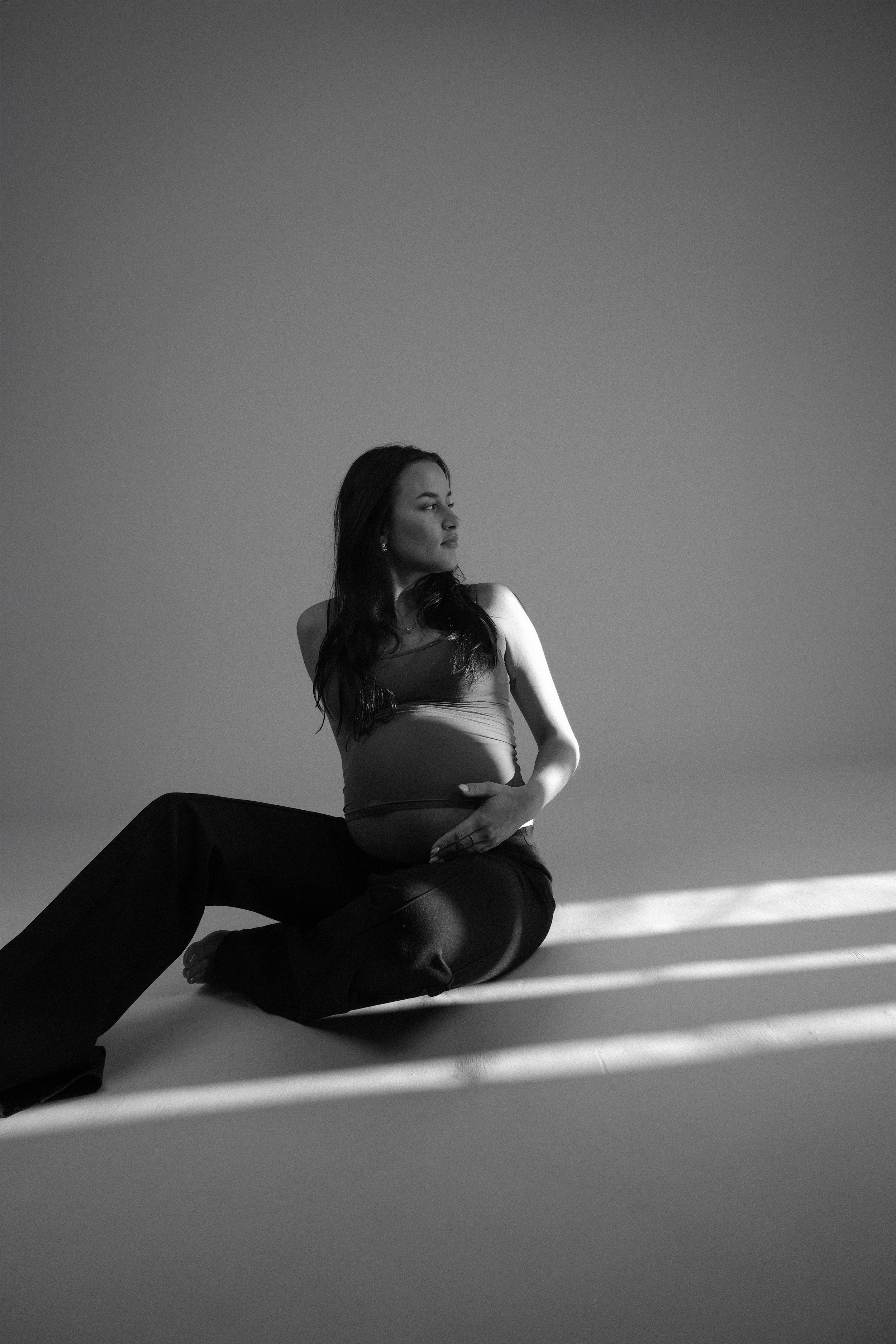 A pregnant woman sitting on the floor, looking to her left, with photography studio lighting casting diagonal shadows across the scene. The photo is in black and white.