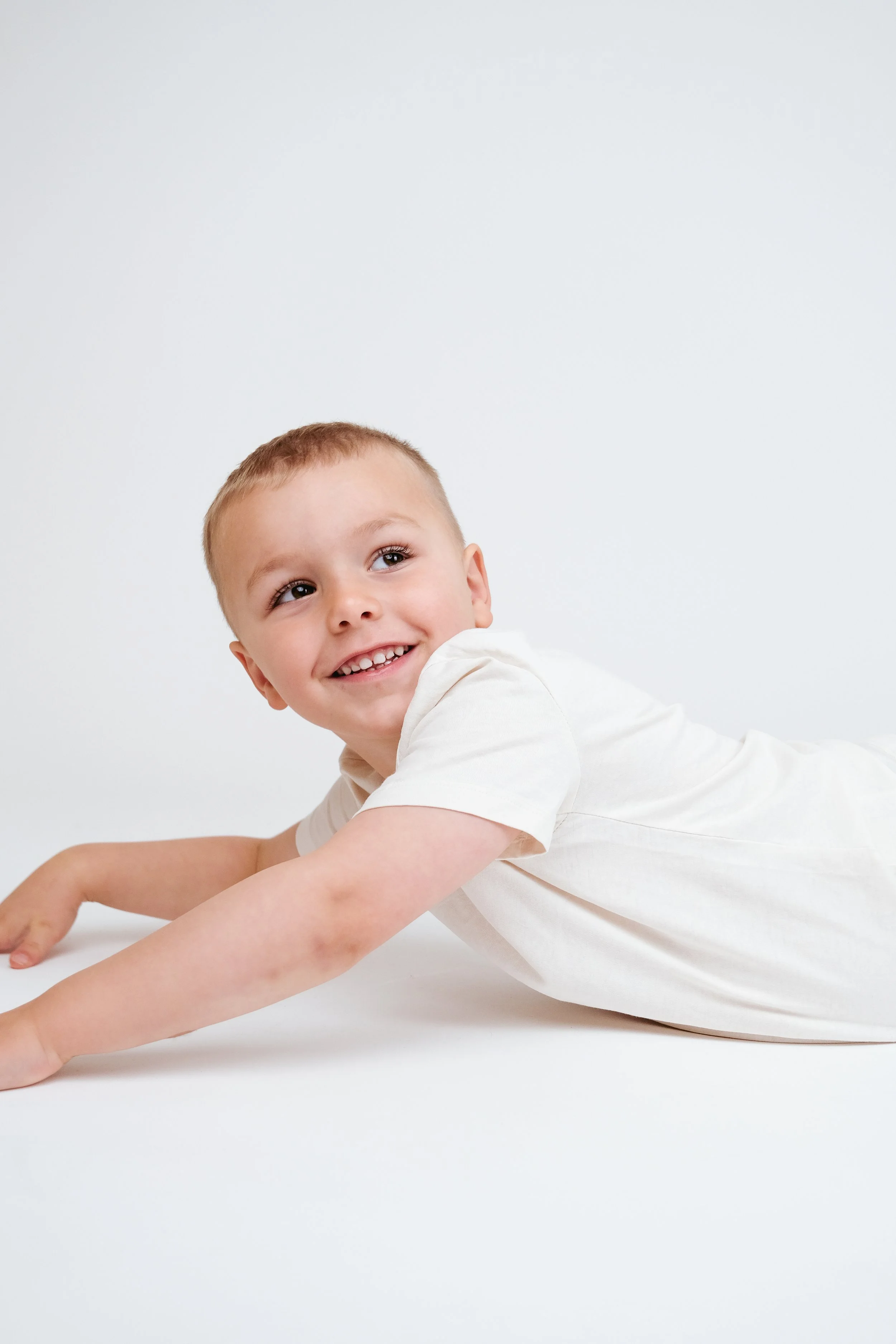 A young boy with short light brown hair, wearing a white shirt, lying on his stomach on a white surface, smiling and looking slightly to the left.