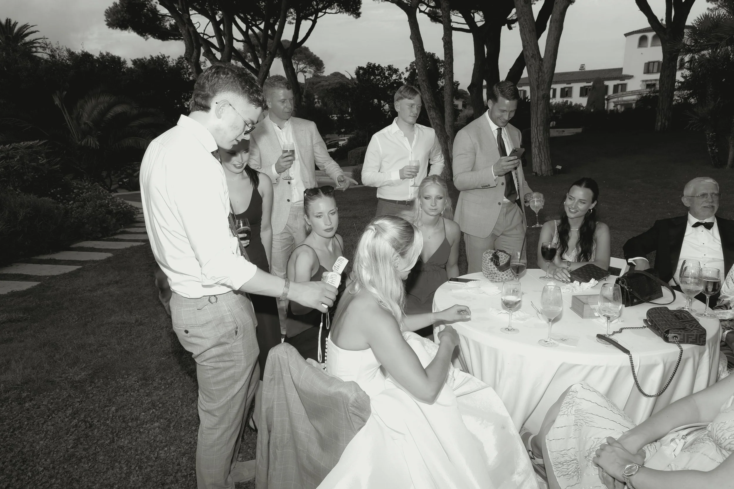 People gathered around a table at an outdoor formal event, some standing and some seated, with drinks and personal belongings on the table.