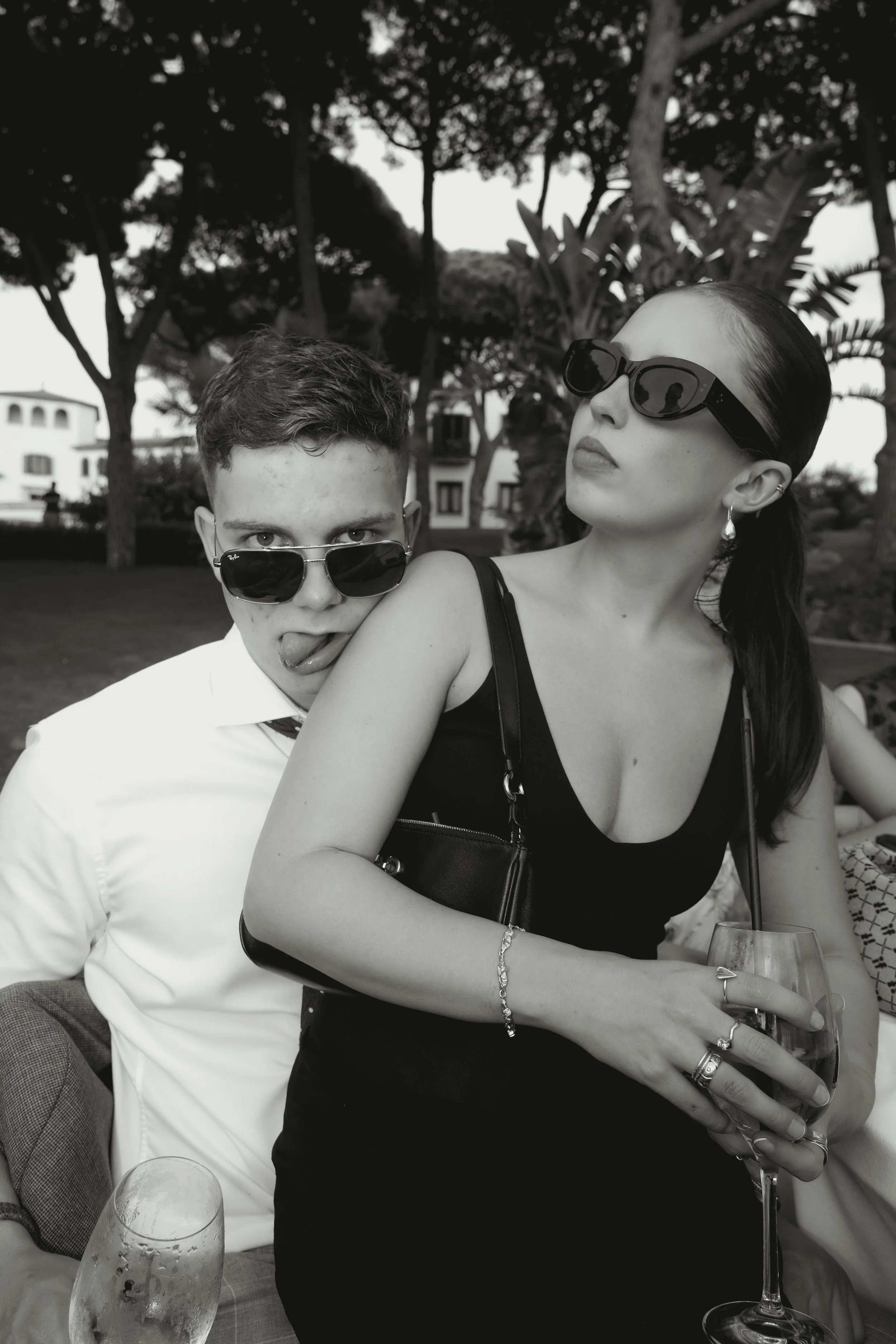 Two people wearing sunglasses, a young man sticking his tongue out and a woman holding a drink, posing closely outdoors with trees and buildings in the background.