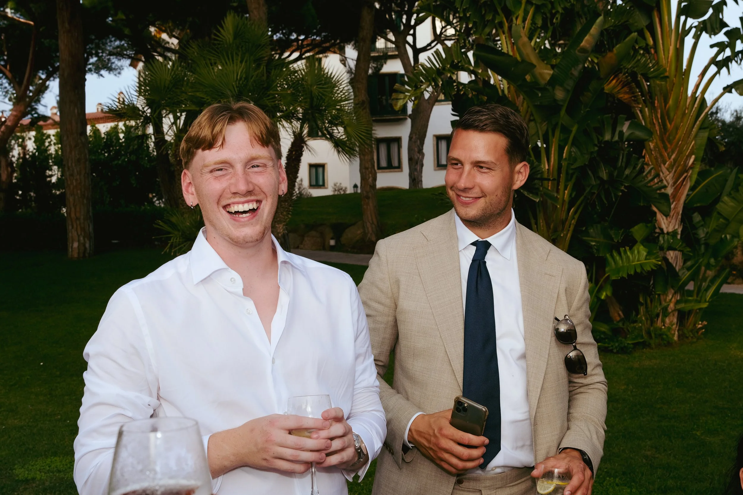 Two men in formal attire are enjoying an outdoor gathering, smiling and holding drinks, with lush greenery and trees in the background.