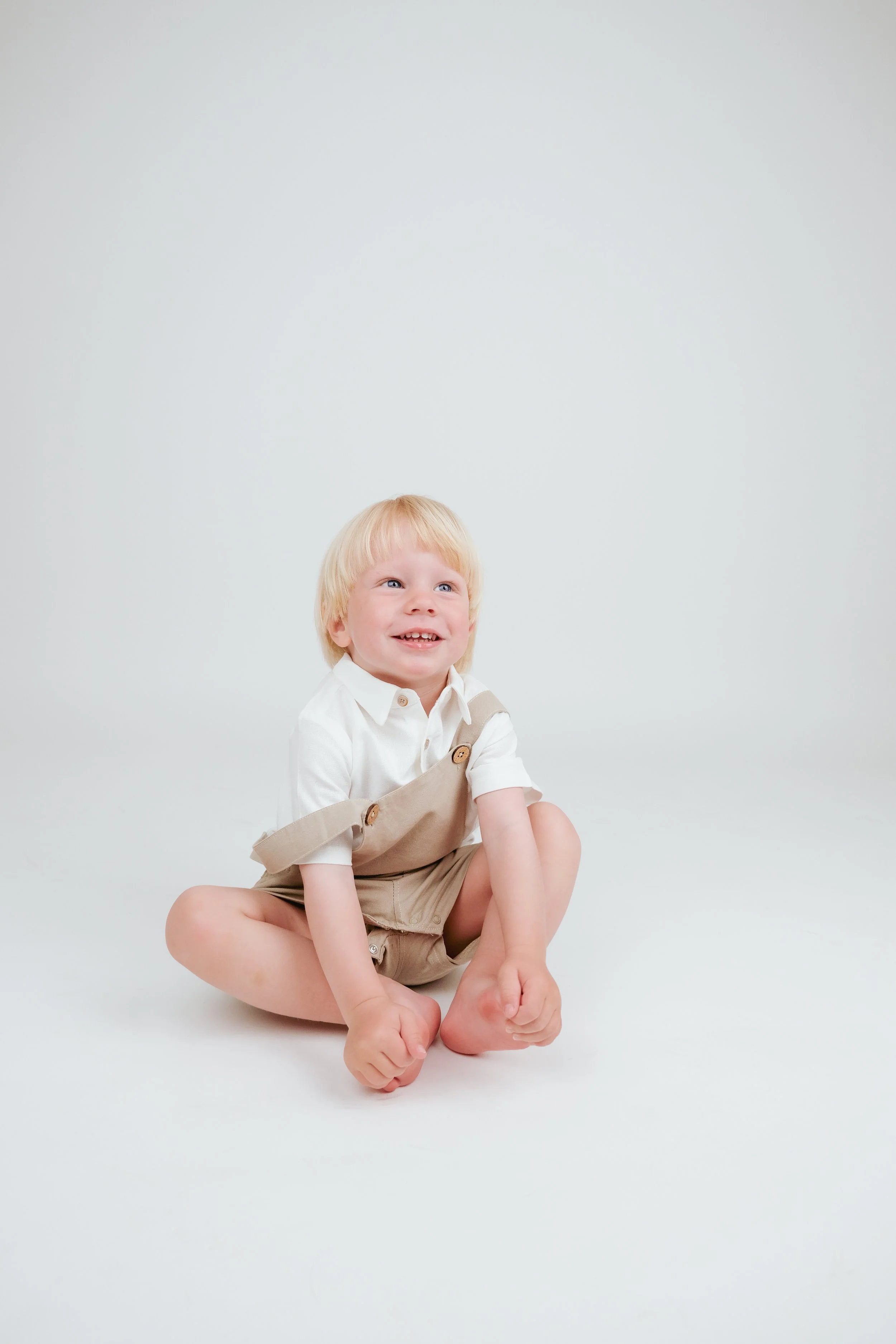A young blonde boy sitting on the white floor, smiling and looking up. He's wearing a white shirt and beige overalls.