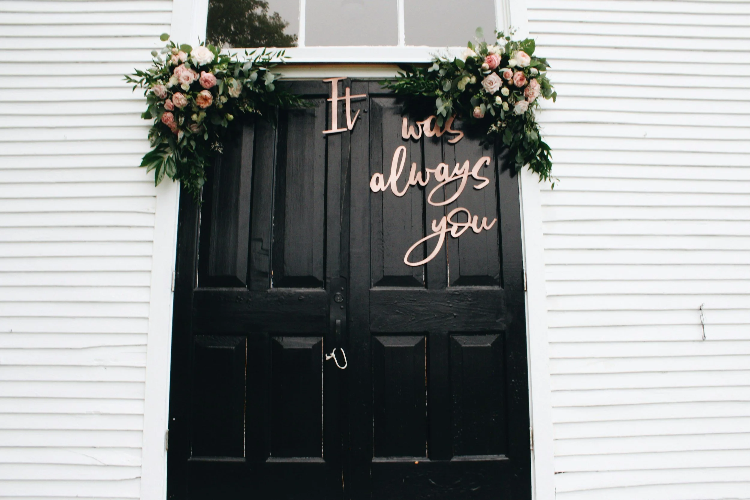 Black double doors decorated with pink and white flowers at the top, featuring a pink script sign that reads 'It was always you'