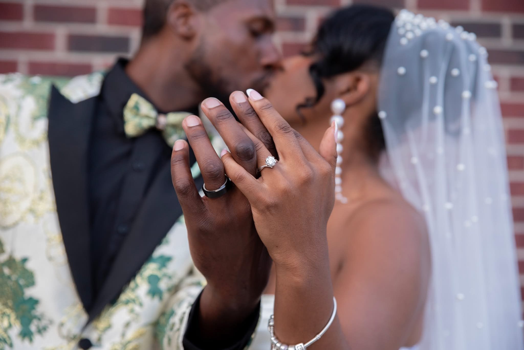 Close-up of a couple holding hands with wedding rings, kissing in the background, bride wearing a veil and earrings, groom wearing a suit with a floral pattern and bow tie, brick wall in the background.