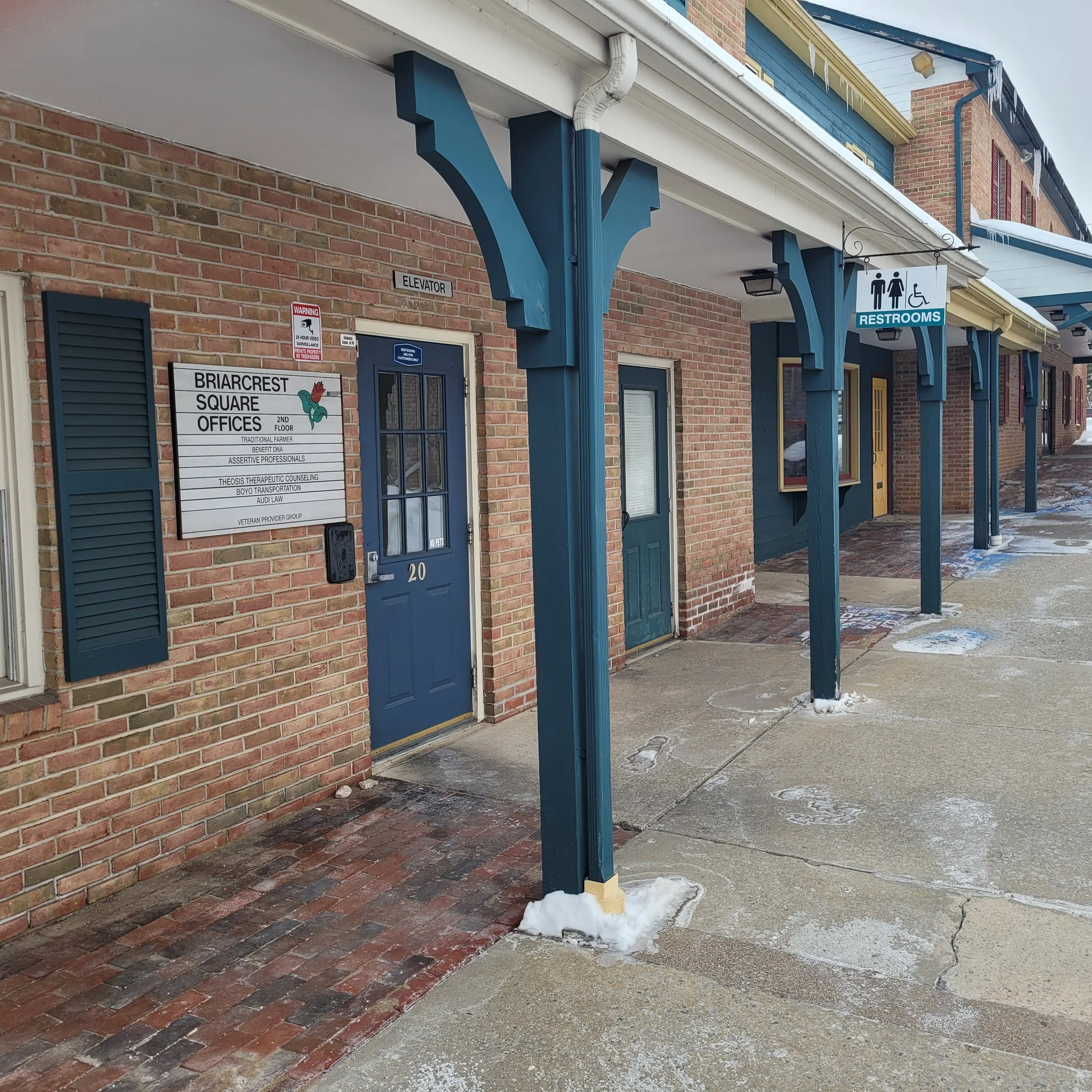 Brick building with blue doors and a covered walkway supported by blue columns. A sign indicates restrooms. Snow and footprints are on the sidewalk.