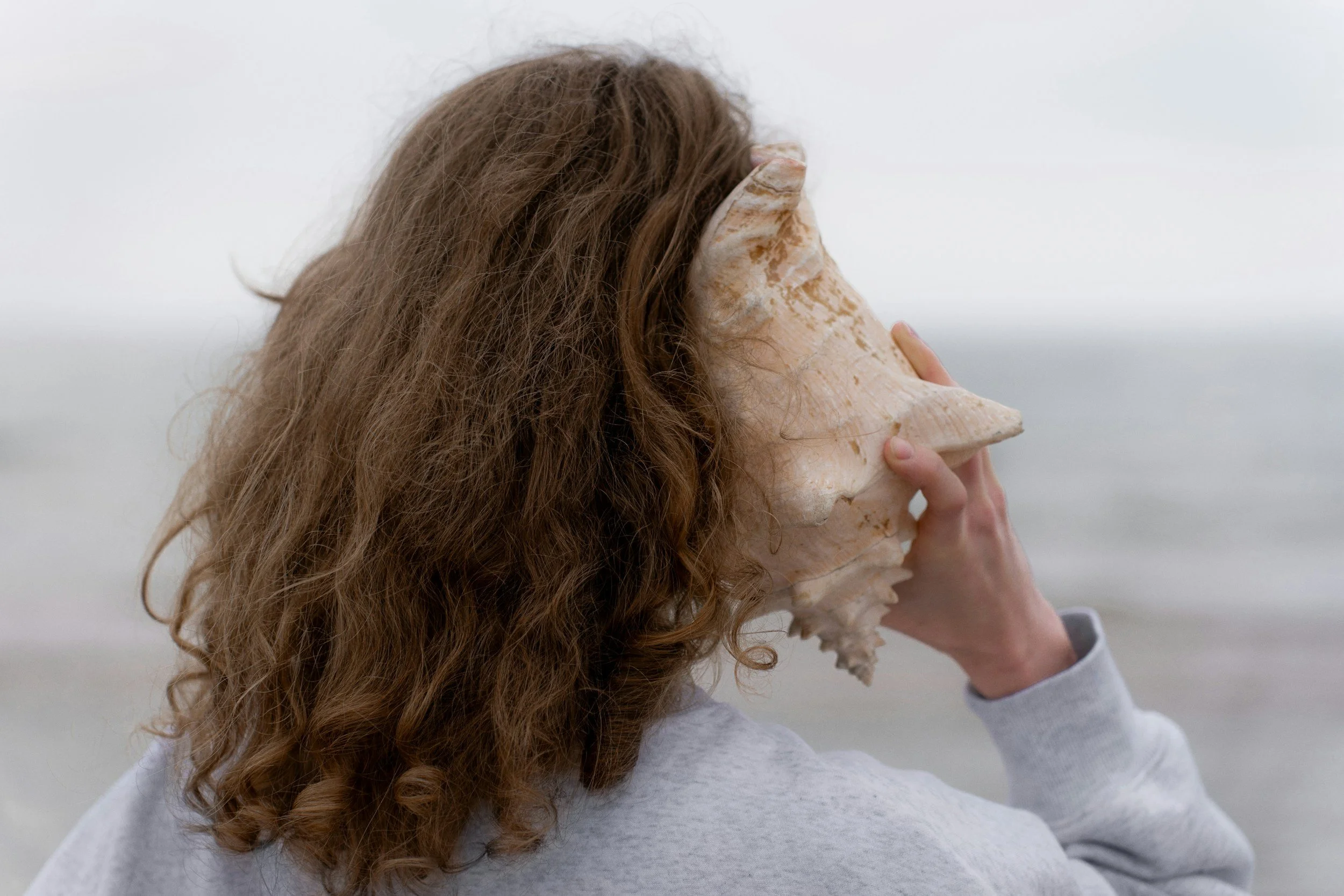 Person with curly brown hair holding a large seashell in front of their face at the beach.