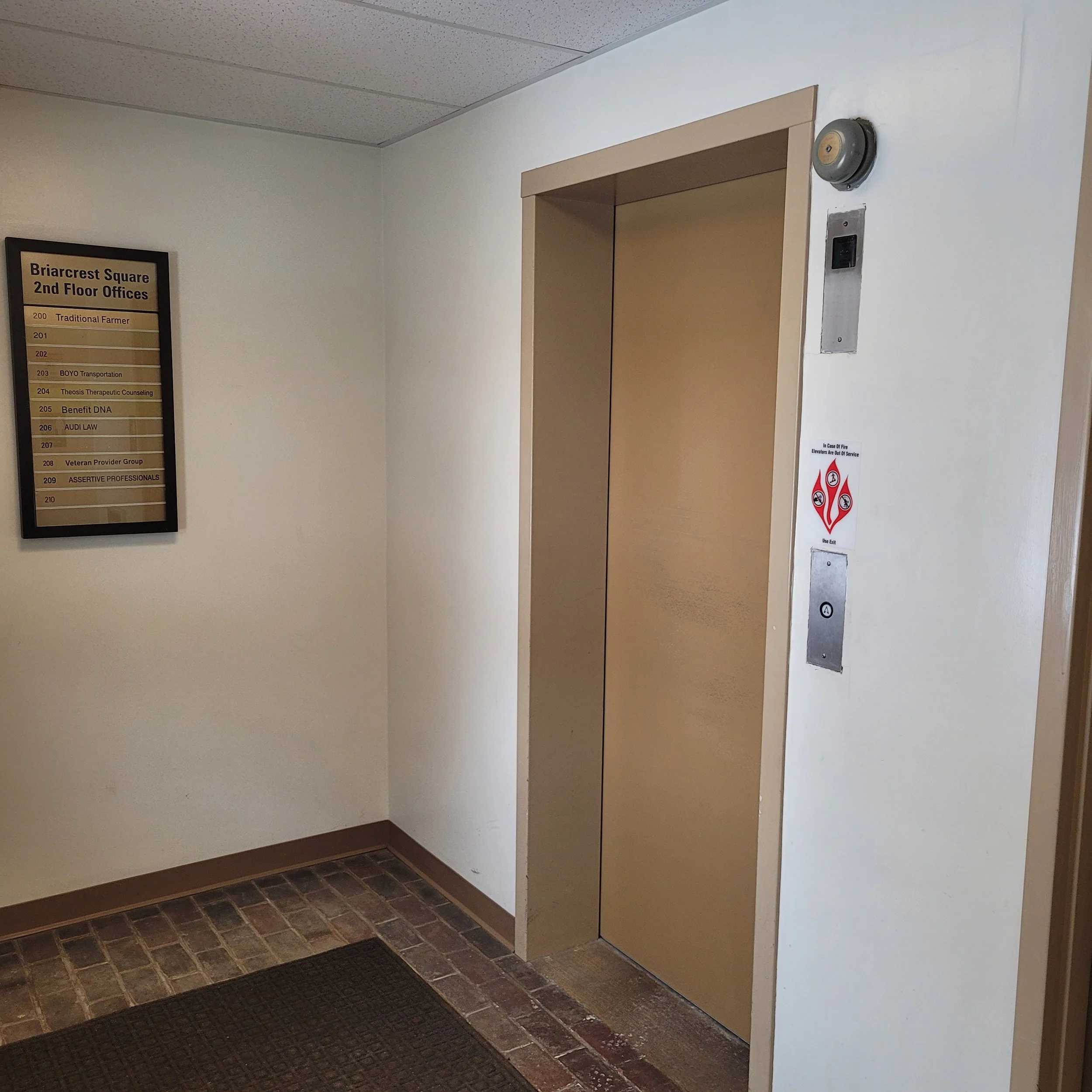 Elevator doors in a building hallway, next to fire safety and elevator buttons, with a directory sign on the wall listing offices on the second floor.